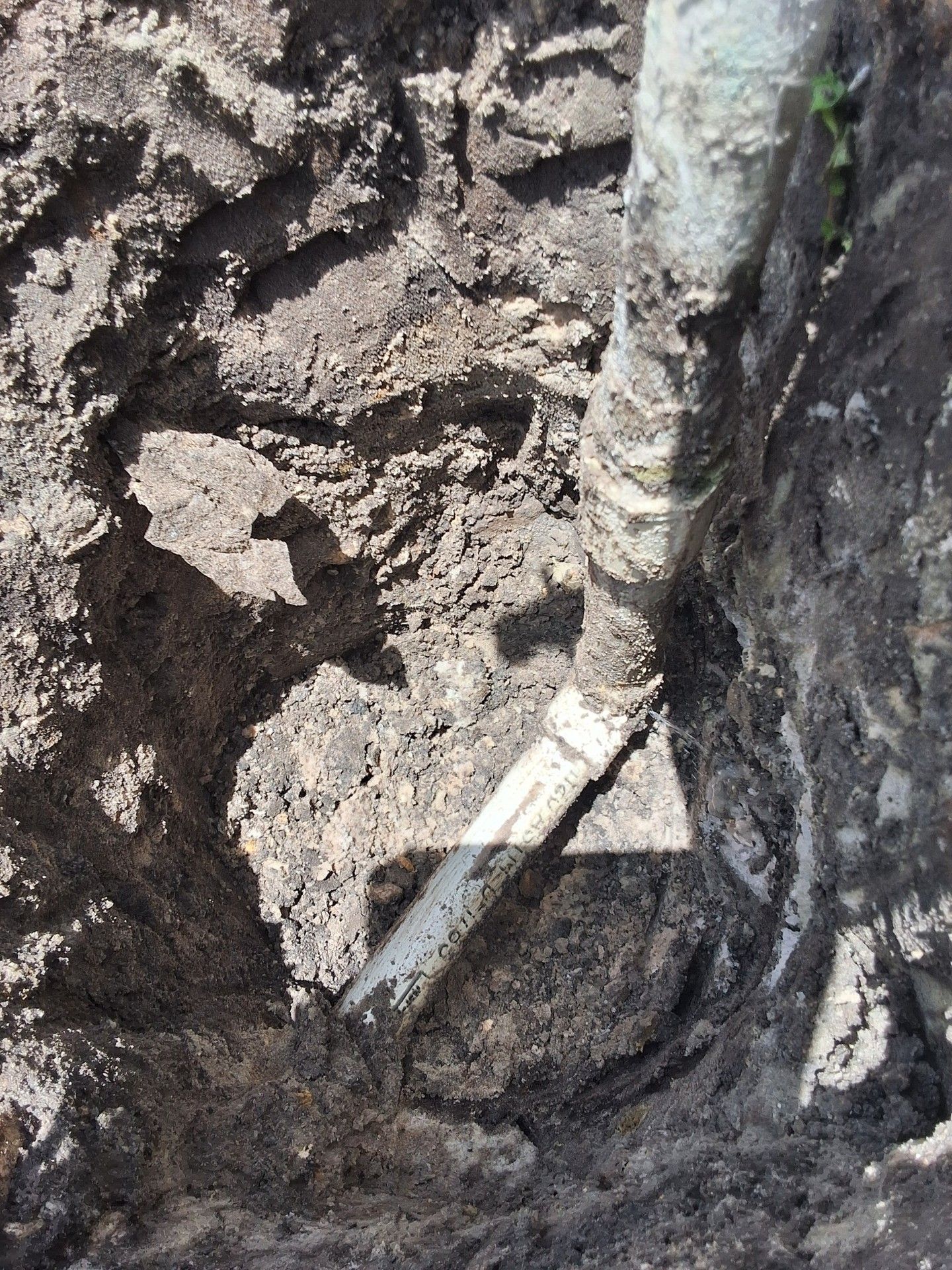 Shovel digging into dark soil beside a white pipe in a muddy trench