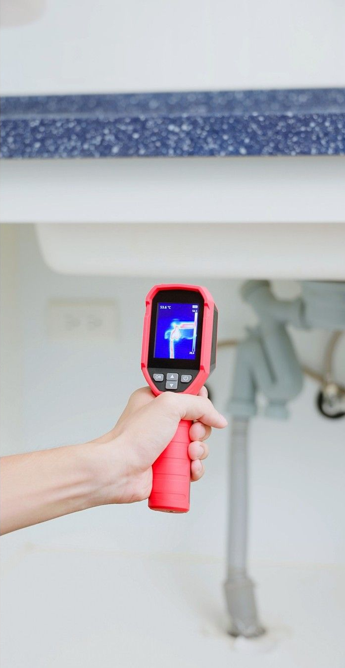 Handheld thermal camera aimed at an under-sink pipe and cabinet underside for inspection