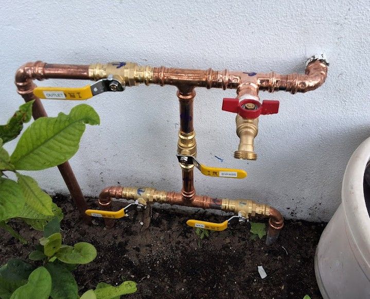 Copper pipe irrigation manifold with yellow valves and a red shutoff, beside a plant and garden bed.