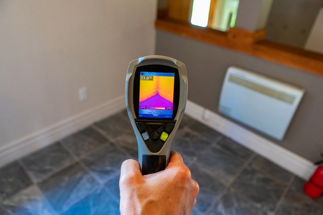 Handheld thermal camera aimed at a room corner, showing a heat map on the screen