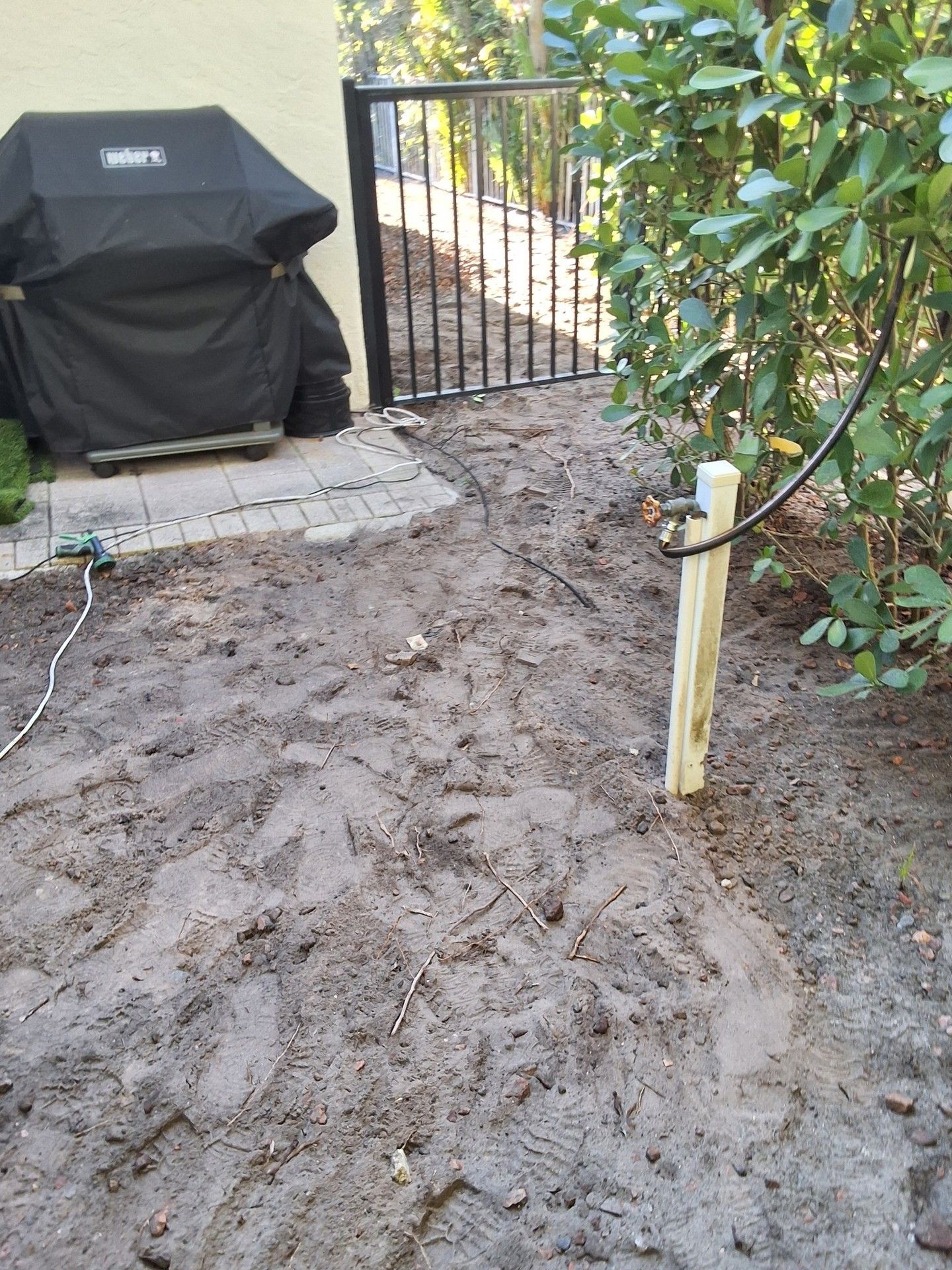 Muddy backyard with a covered grill, gate, and a tree near a tiled patio.