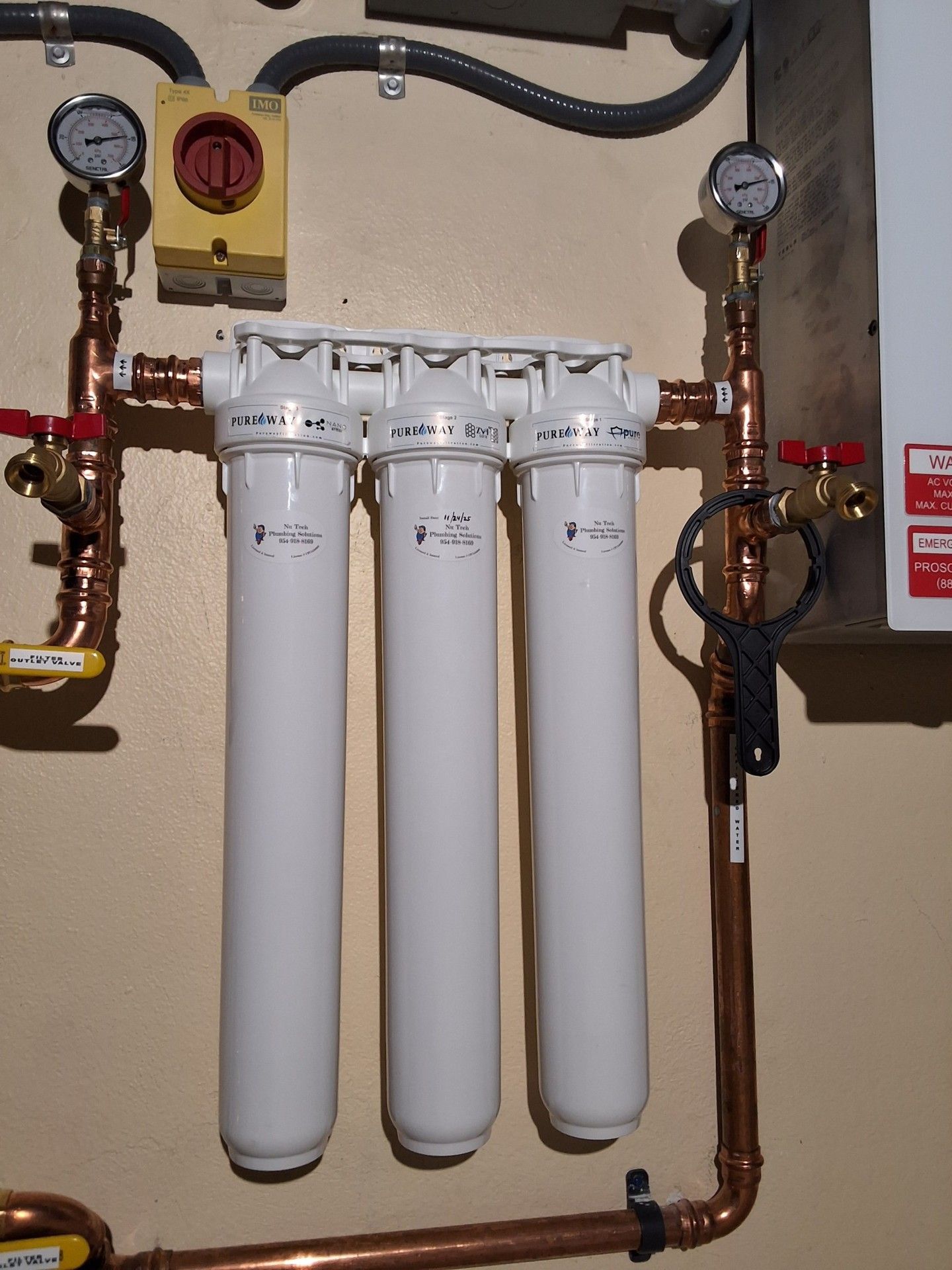 Three white water filters mounted on copper pipes with gauges in a utility room.