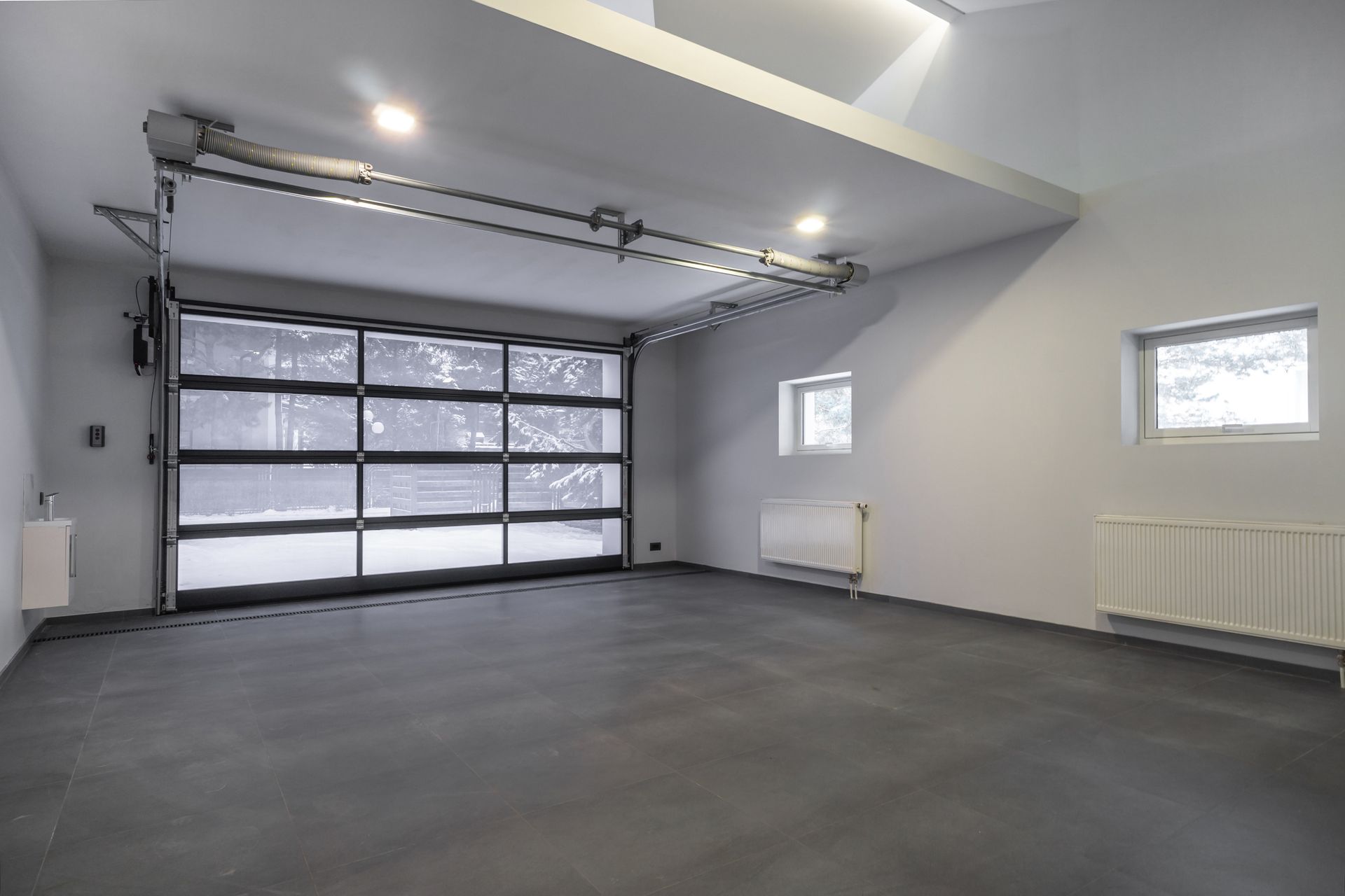 Empty modern garage with gray floor, white walls, large glass overhead door, and two small windows