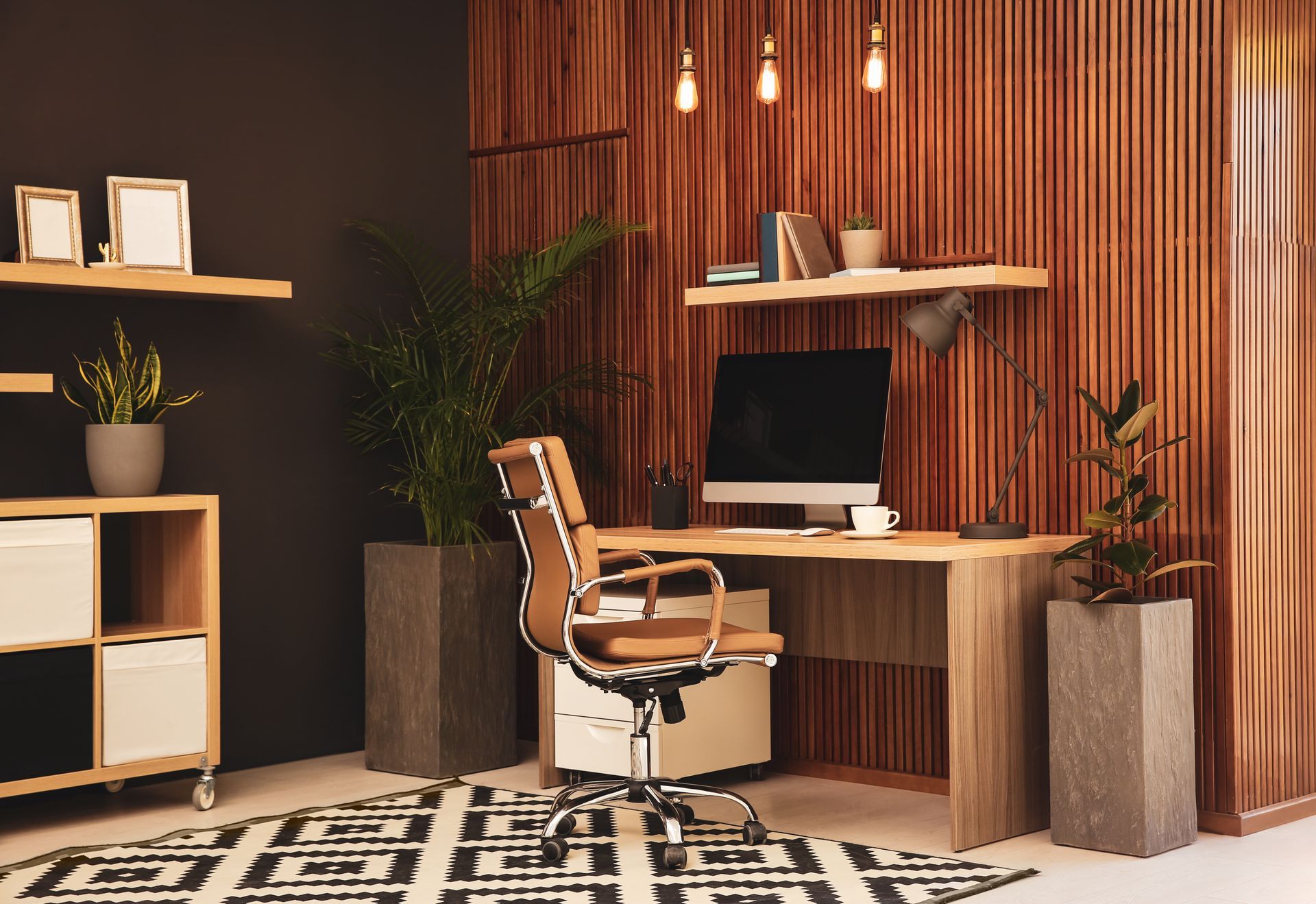 Modern home office with wooden desk, black monitor, tan chair, shelves, plants, and patterned rug