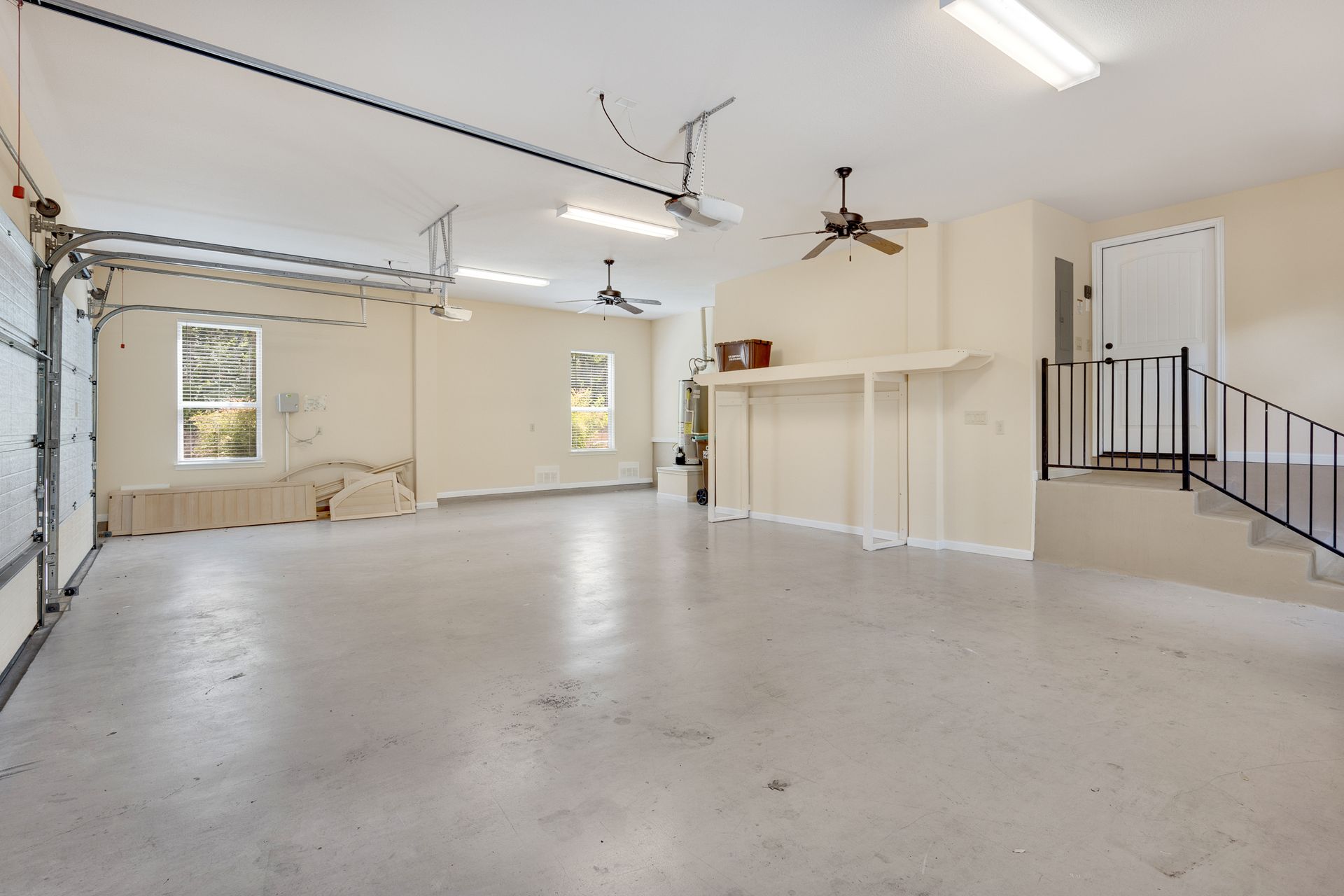 Empty garage with white walls, concrete floor, overhead lights, windows, and a staircase to the right.