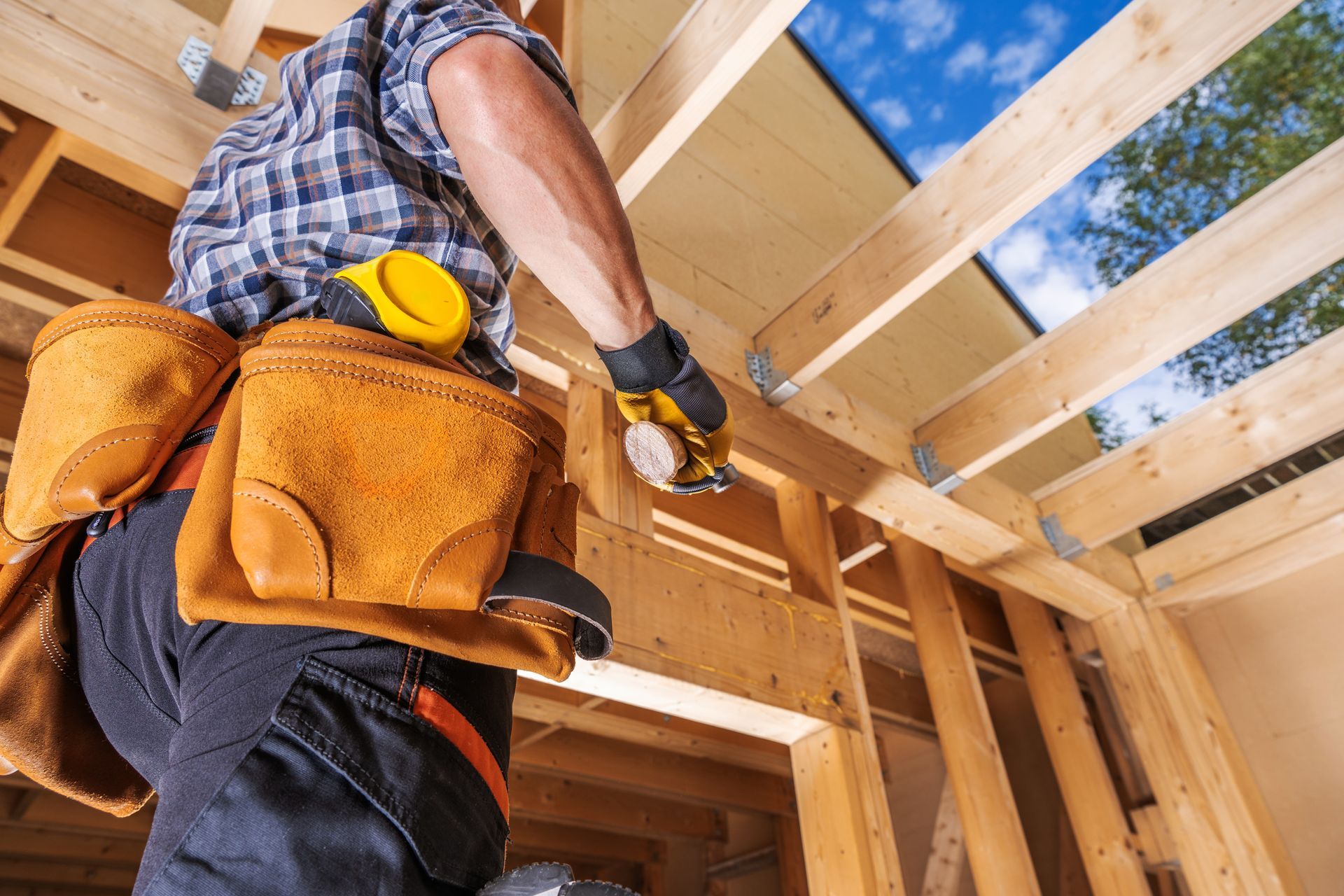 Construction worker installing ceiling beams in a wooden frame building, wearing a tool belt and gloves.