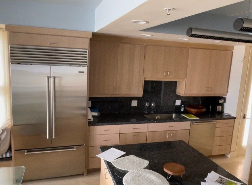 Modern kitchen with stainless fridge, light wood cabinets, black countertops, and an island.