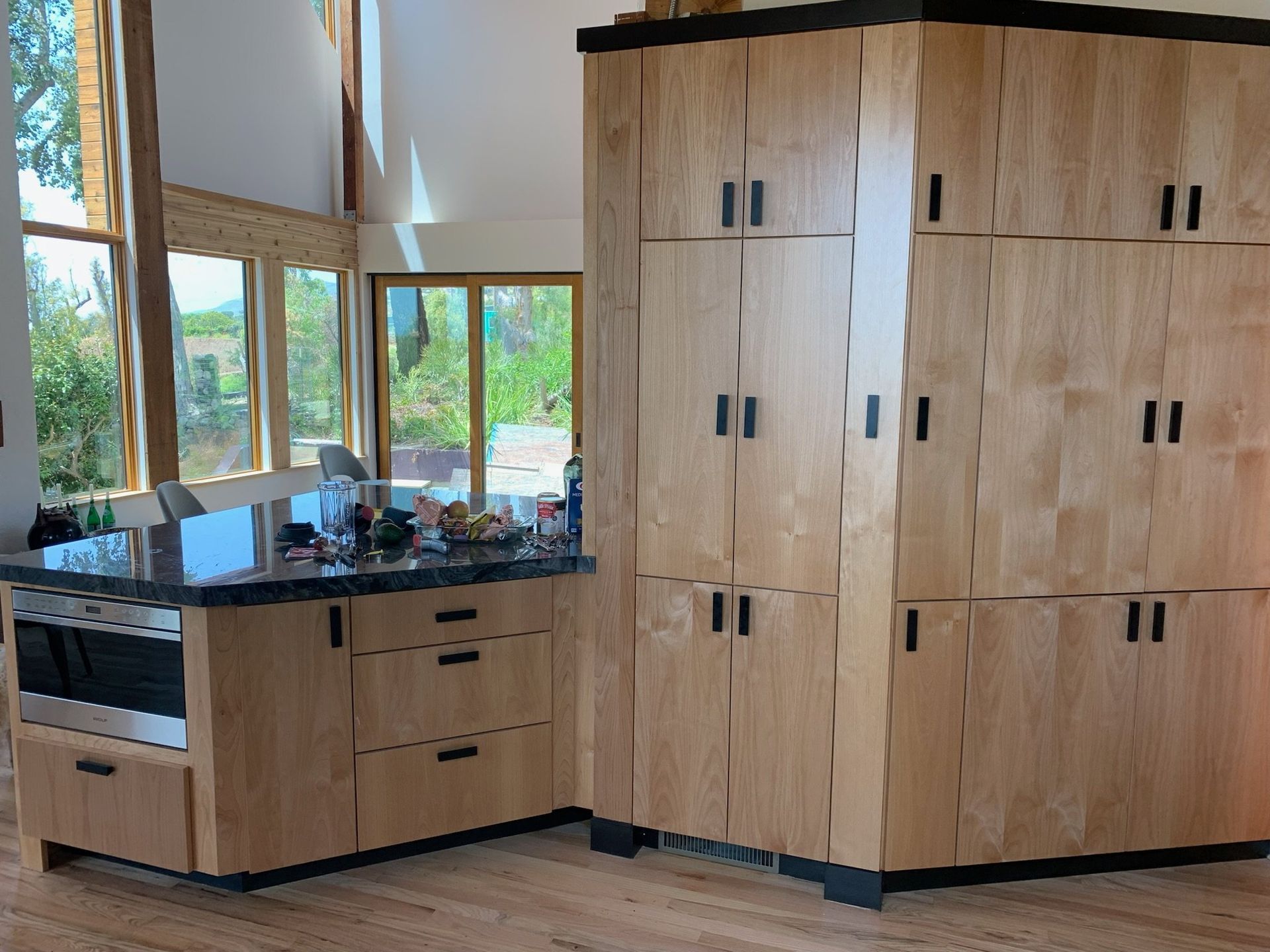 Modern kitchen with wood cabinets, black countertop, and large windows overlooking greenery