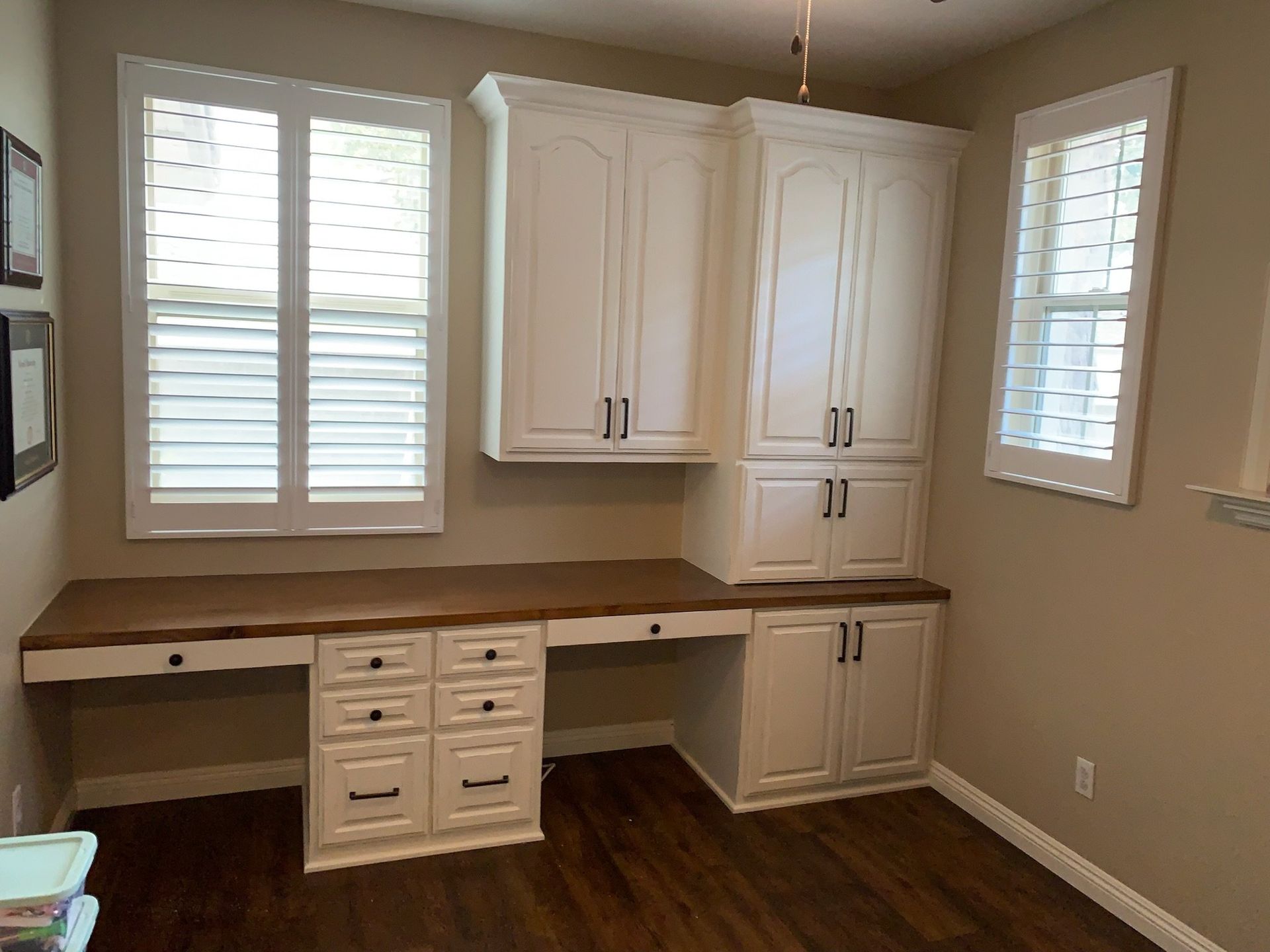 Built-in white cabinets and desk in a beige home office with windows and dark wood floor