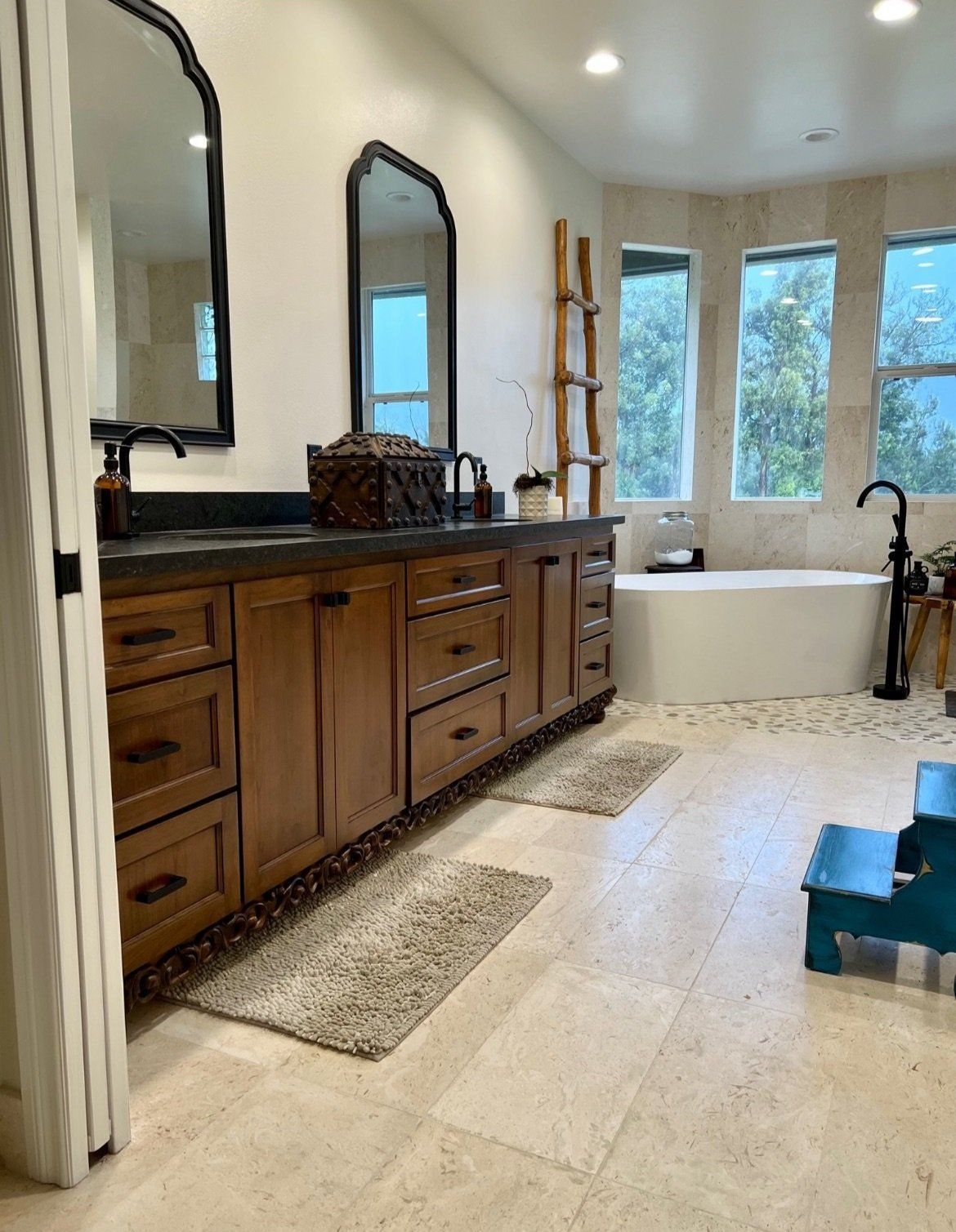 Bright bathroom with wooden vanity, mirrors, freestanding tub, woven rugs, and large windows.