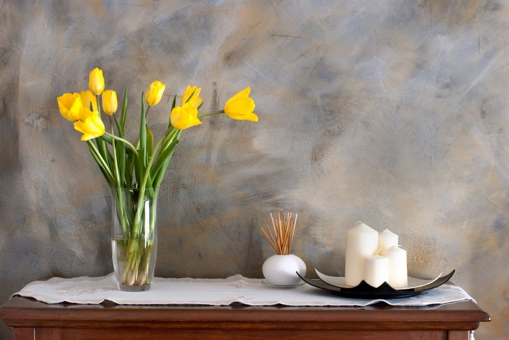 Yellow daffodils in a glass vase beside white candles and a bowl on a wooden table