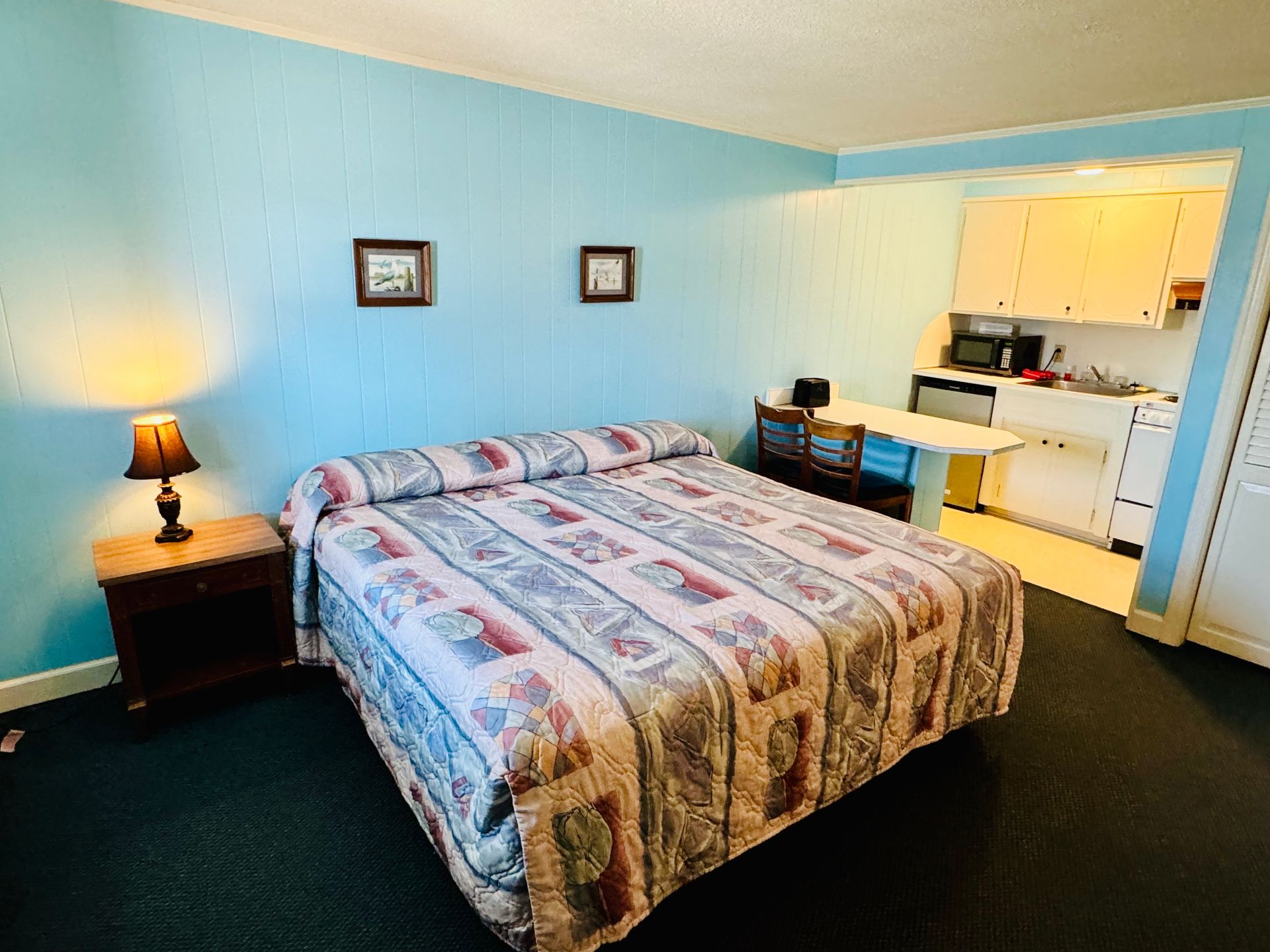 A hotel room with a bed , nightstand , lamp and kitchen.