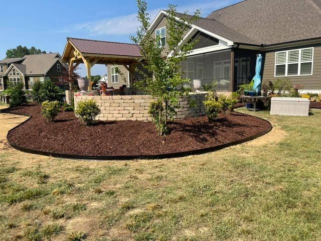 Residential backyard with a curved mulch garden bed, small trees, and a covered patio by a house.