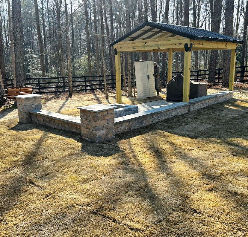 Small outdoor pavilion with a wooden roof and stone columns on a concrete pad in a wooded yard