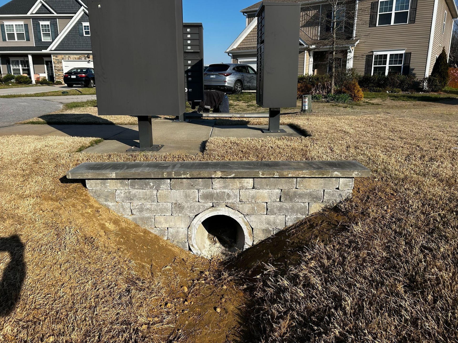 Storm drain culvert under a stone wall in a suburban yard with gravel and houses in the background