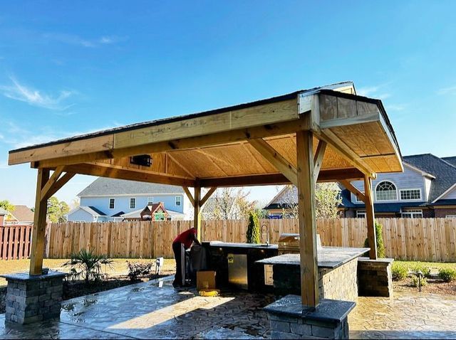 Wooden picnic shelter with a shingled roof in a sunny backyard, with a person working inside.