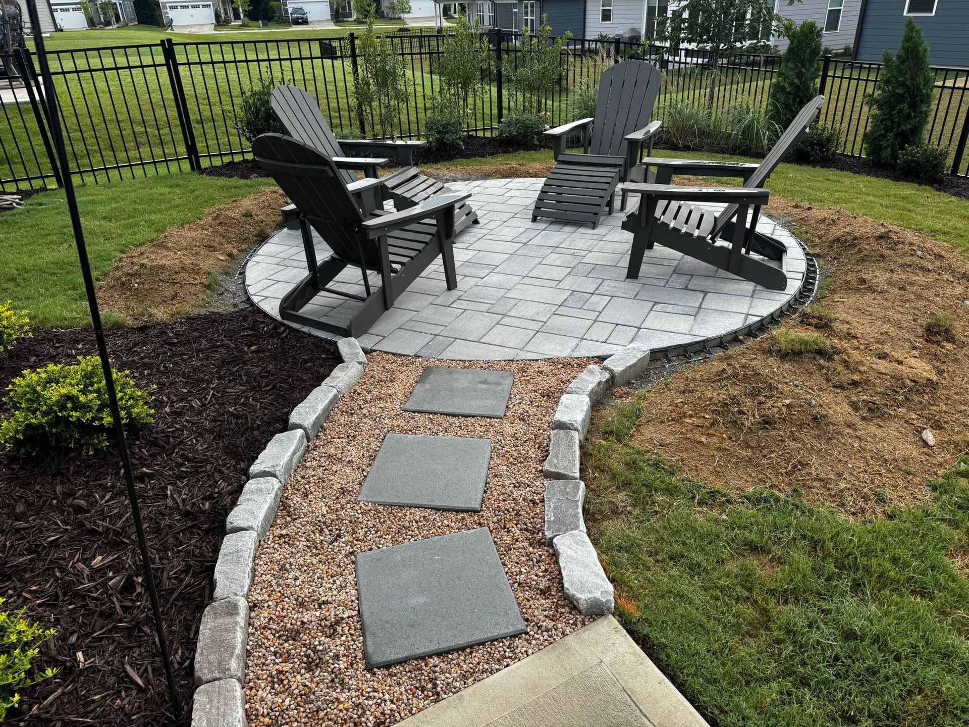 Backyard stone patio with curved paver path and three black Adirondack chairs around a fire pit area