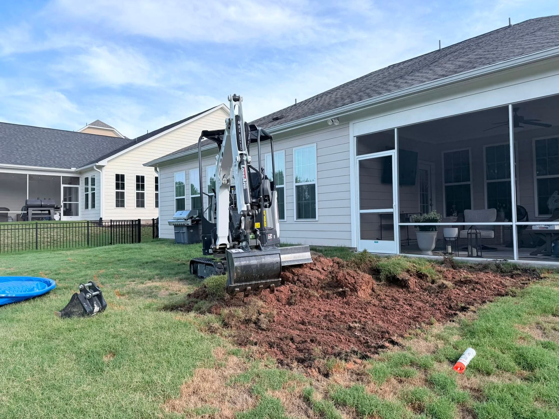 Backhoe digging a large hole in a backyard beside a white house with a screened porch