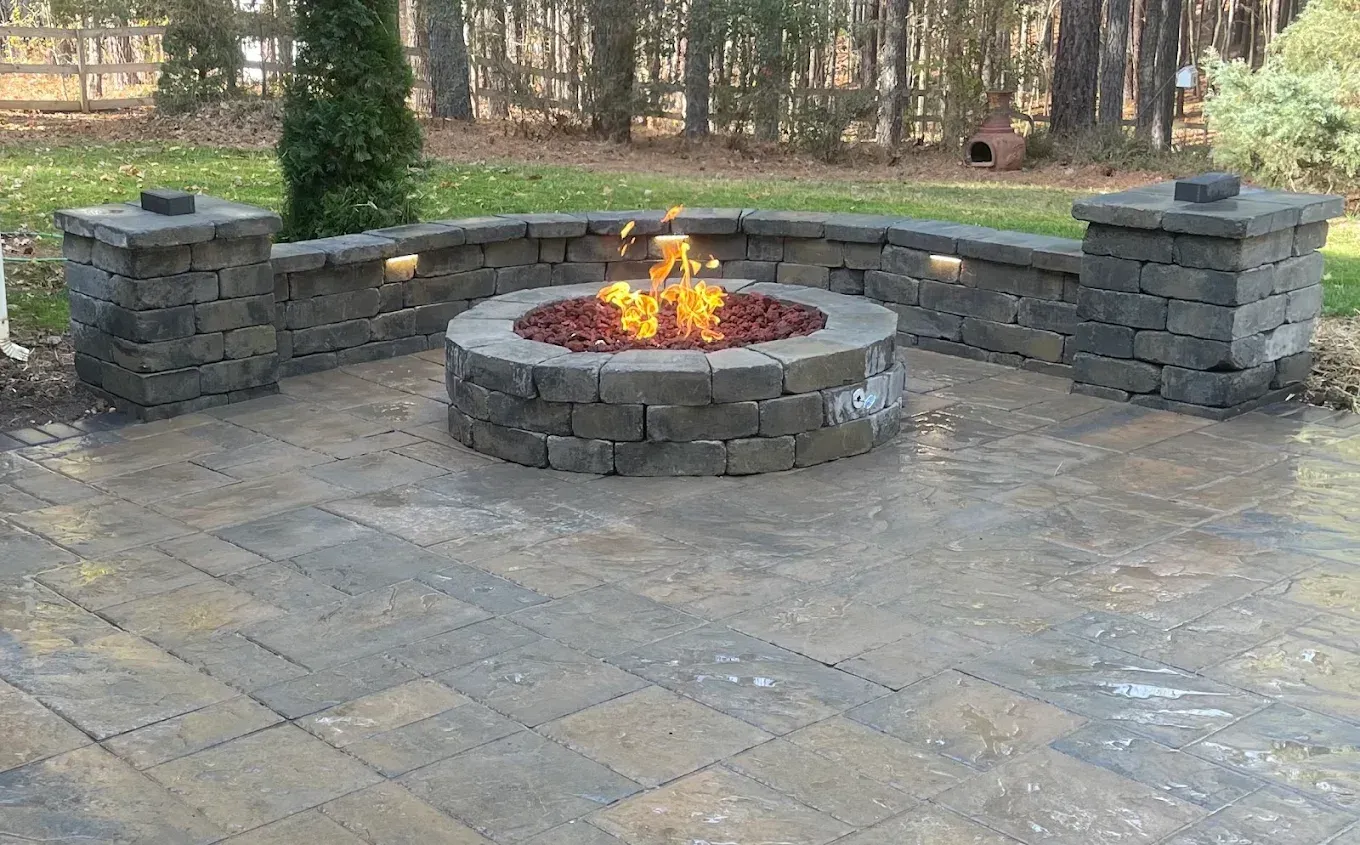 Stone patio with a circular fire pit burning at the center, backed by a curved bench and wooded area.