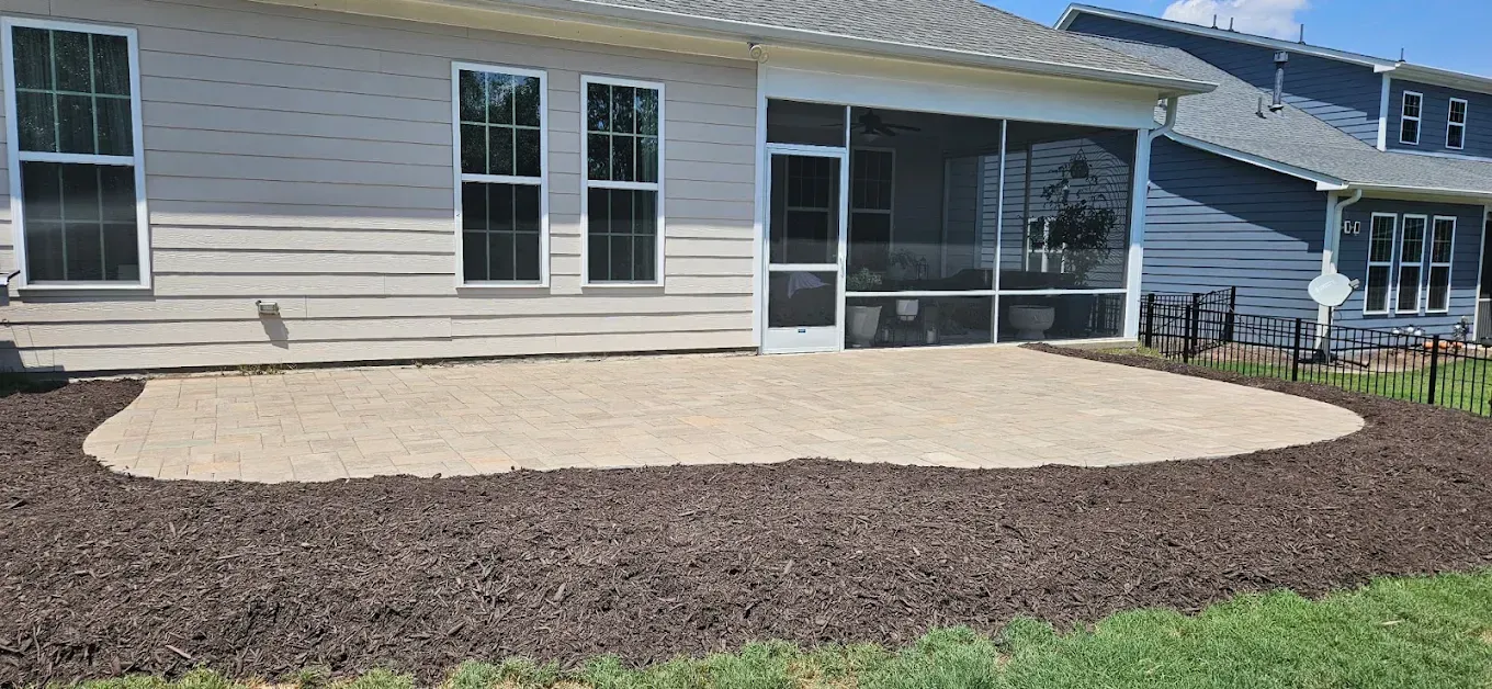 Backyard patio with tan pavers beside a house, bordered by dark mulch and grass