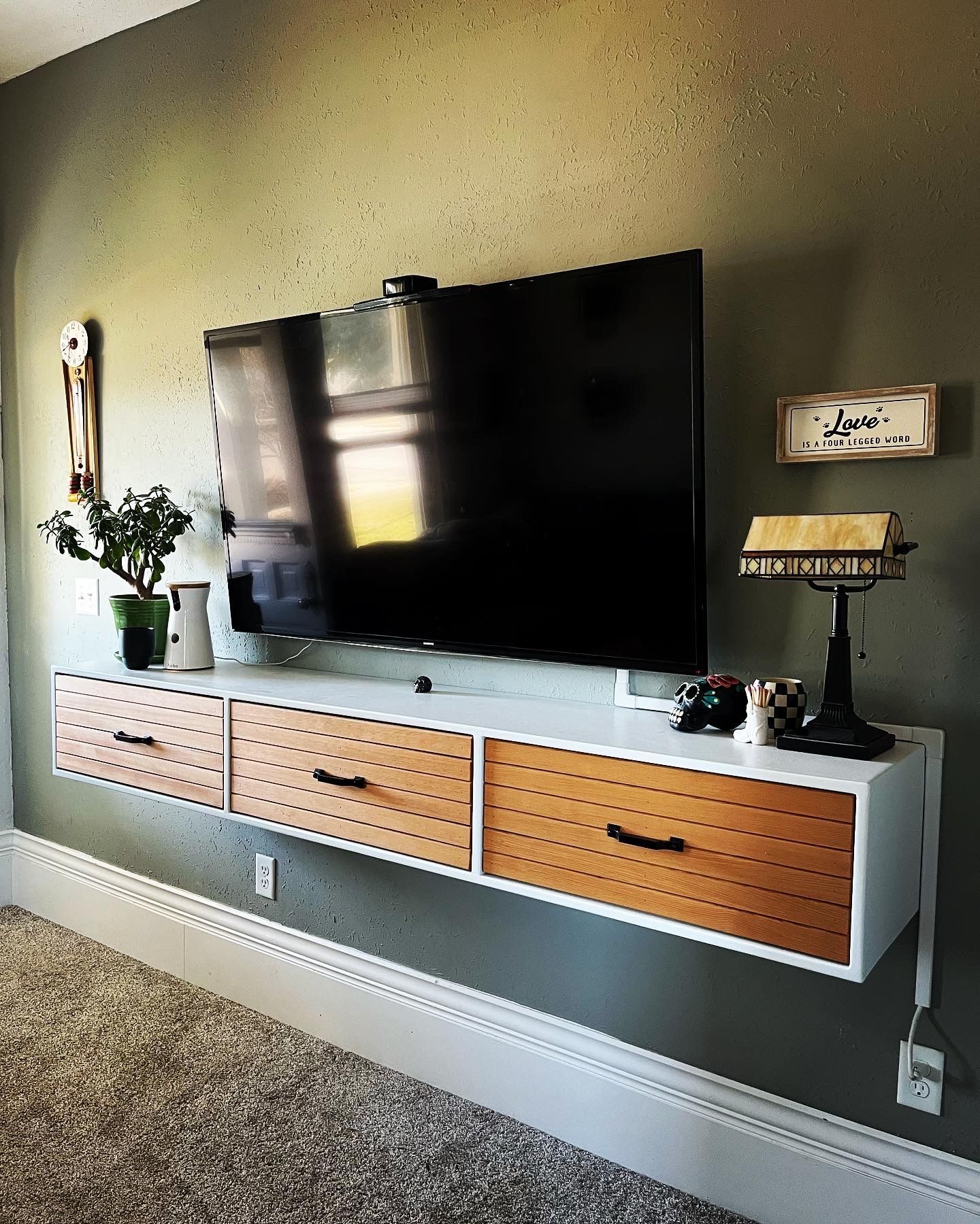 A flat-screen TV mounted above a floating wooden cabinet with three textured drawers against a green wall.