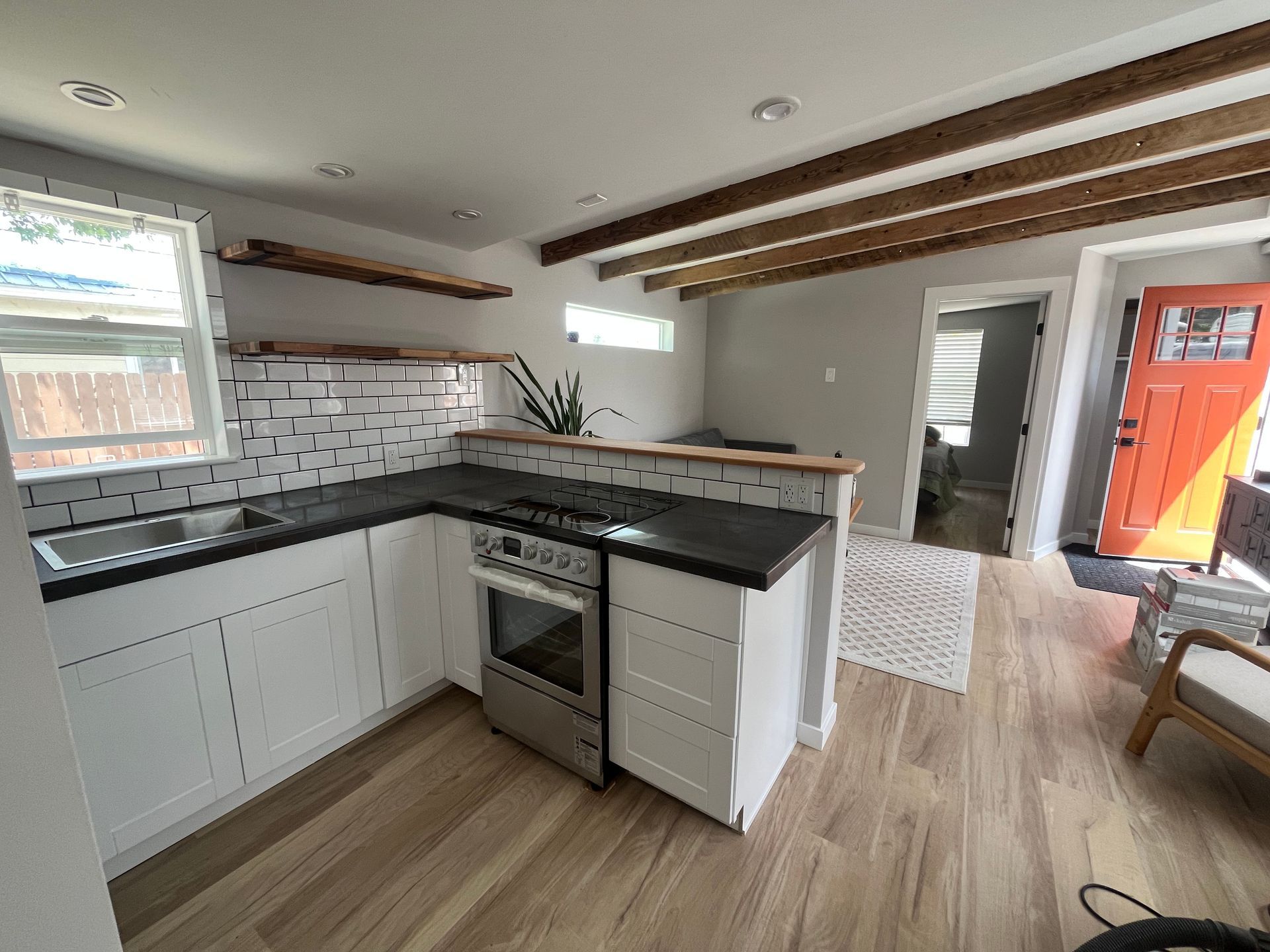 Modern kitchen with white cabinets, black countertops, wood-toned flooring, exposed ceiling beams, and orange front door.
