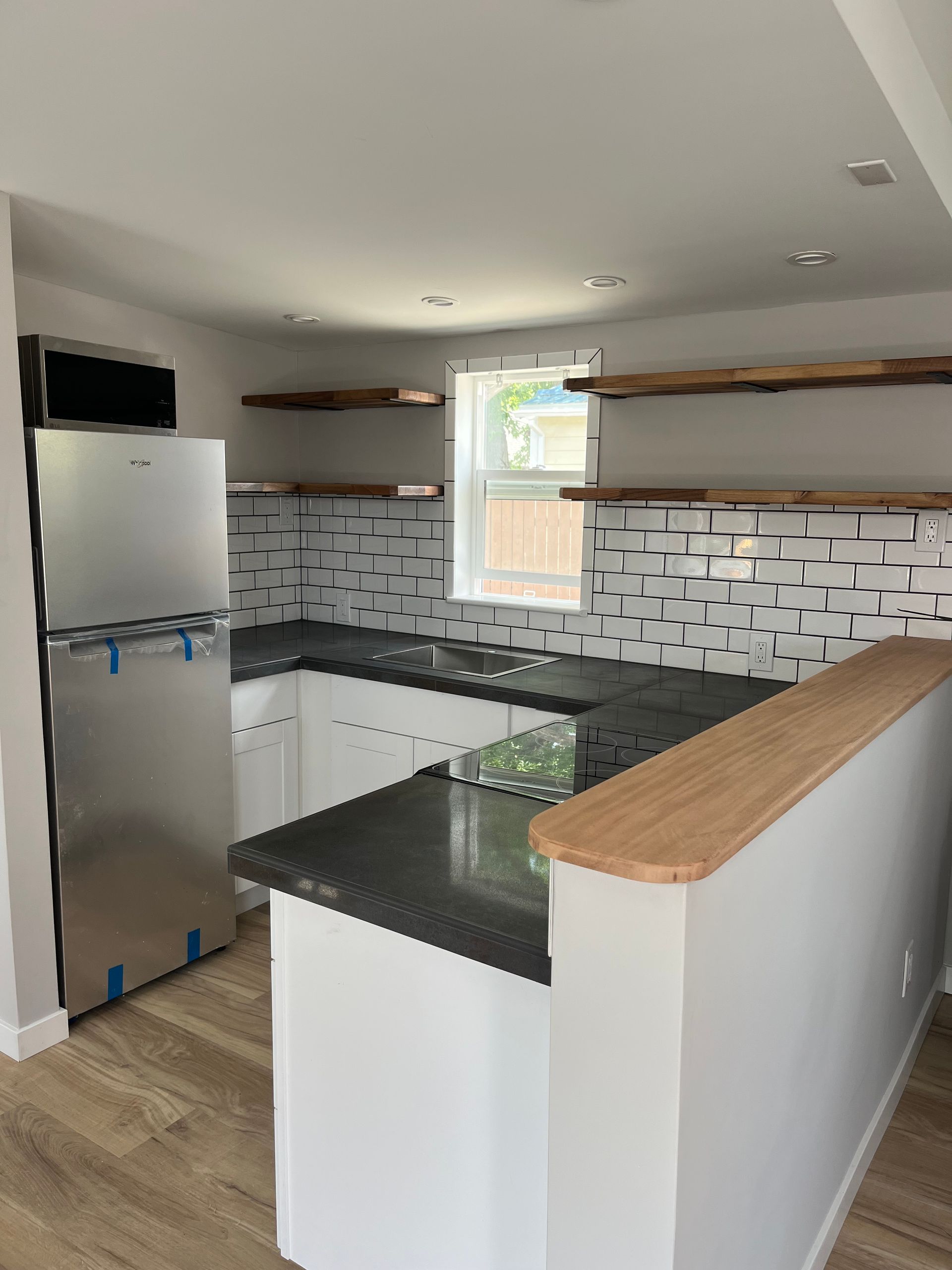 A modern kitchen featuring white cabinets, dark countertops, a tile backsplash, stainless steel fridge, and wood shelves.