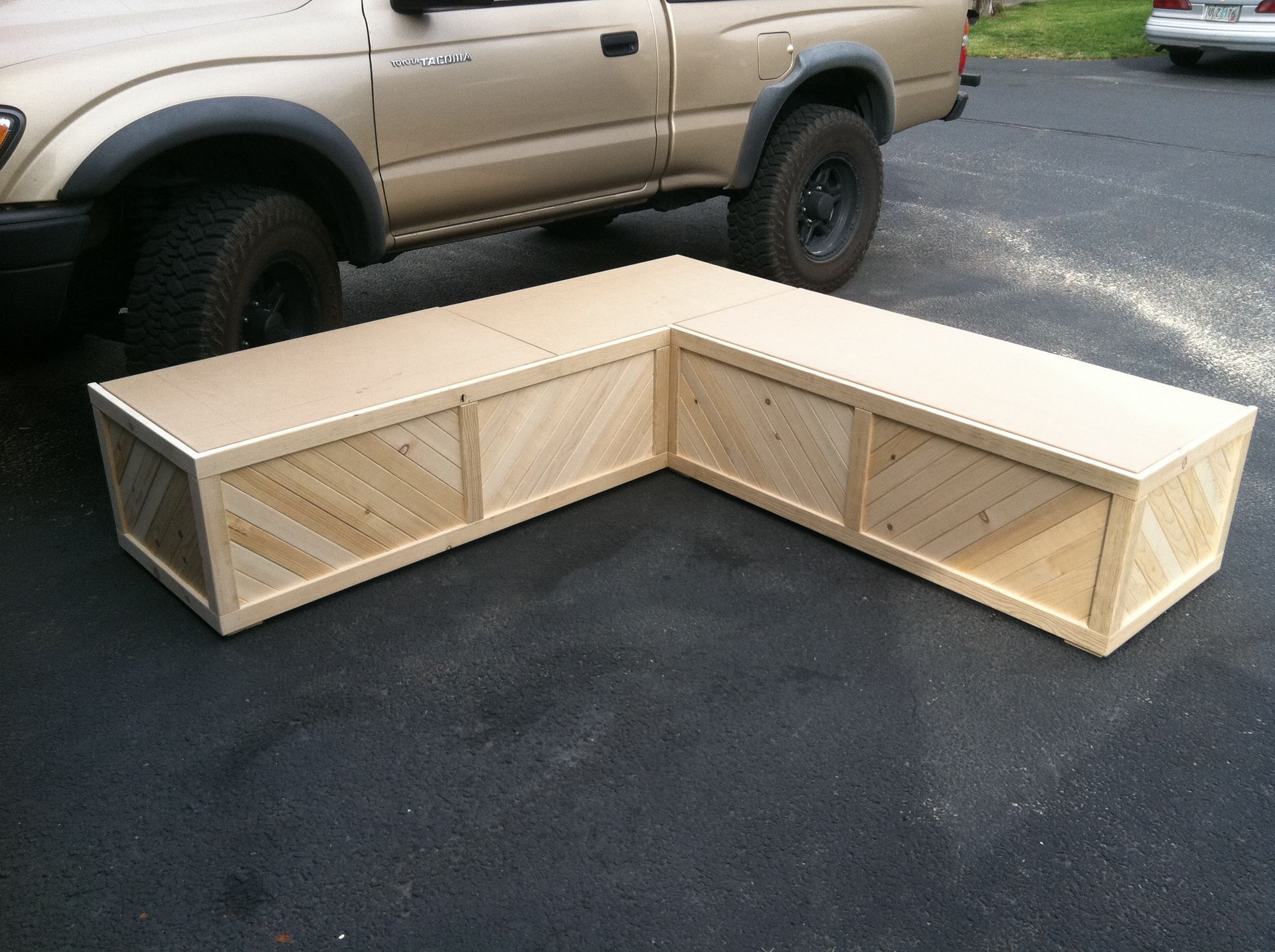 An L-shaped wooden bench with diagonal slat panels, sitting on asphalt next to a tan pickup truck.