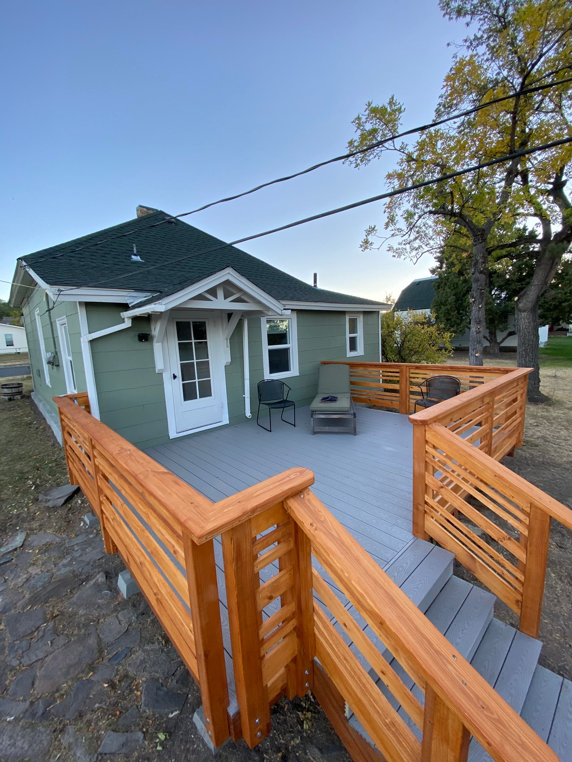 A small green house with a white door and a new wooden deck with gray composite decking in a yard with trees.