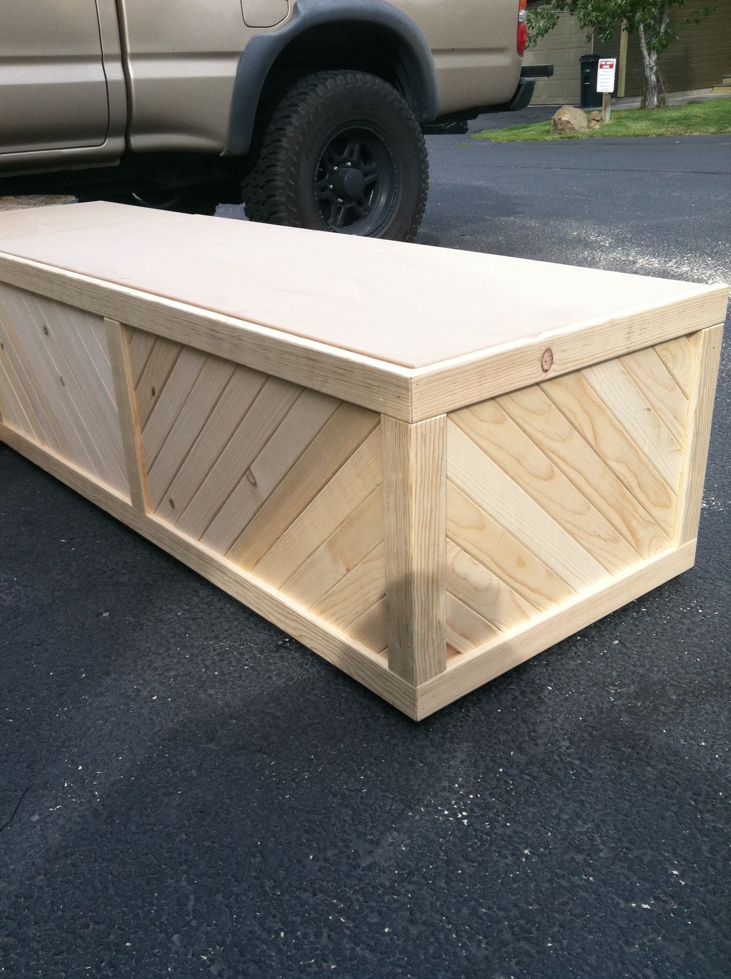 A large wooden storage box with diagonal paneling, resting on pavement near the back of a parked pickup truck.