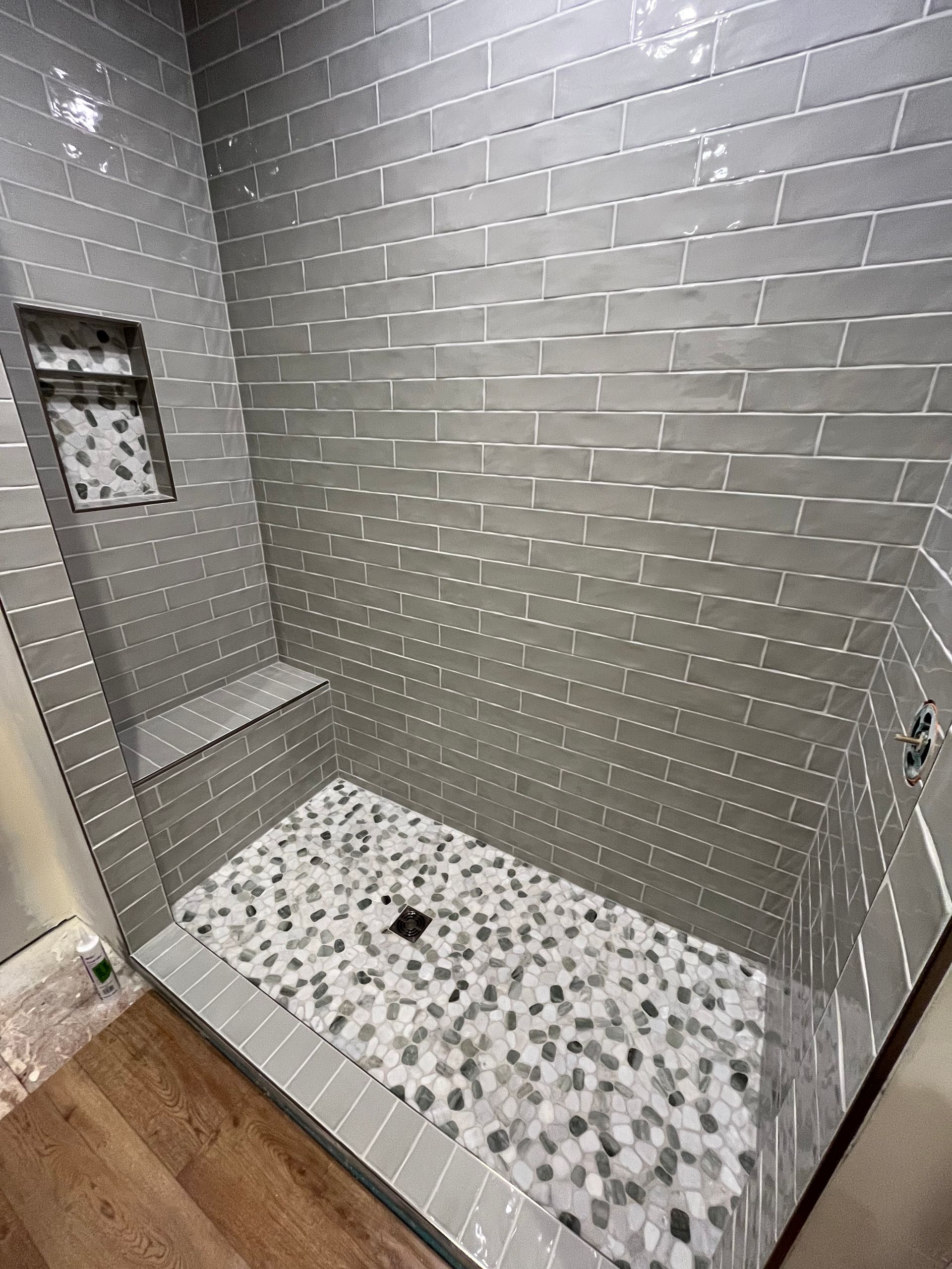 A shower stall with gray subway tile walls, a pebble-tiled floor, a built-in bench, and a small recessed niche.