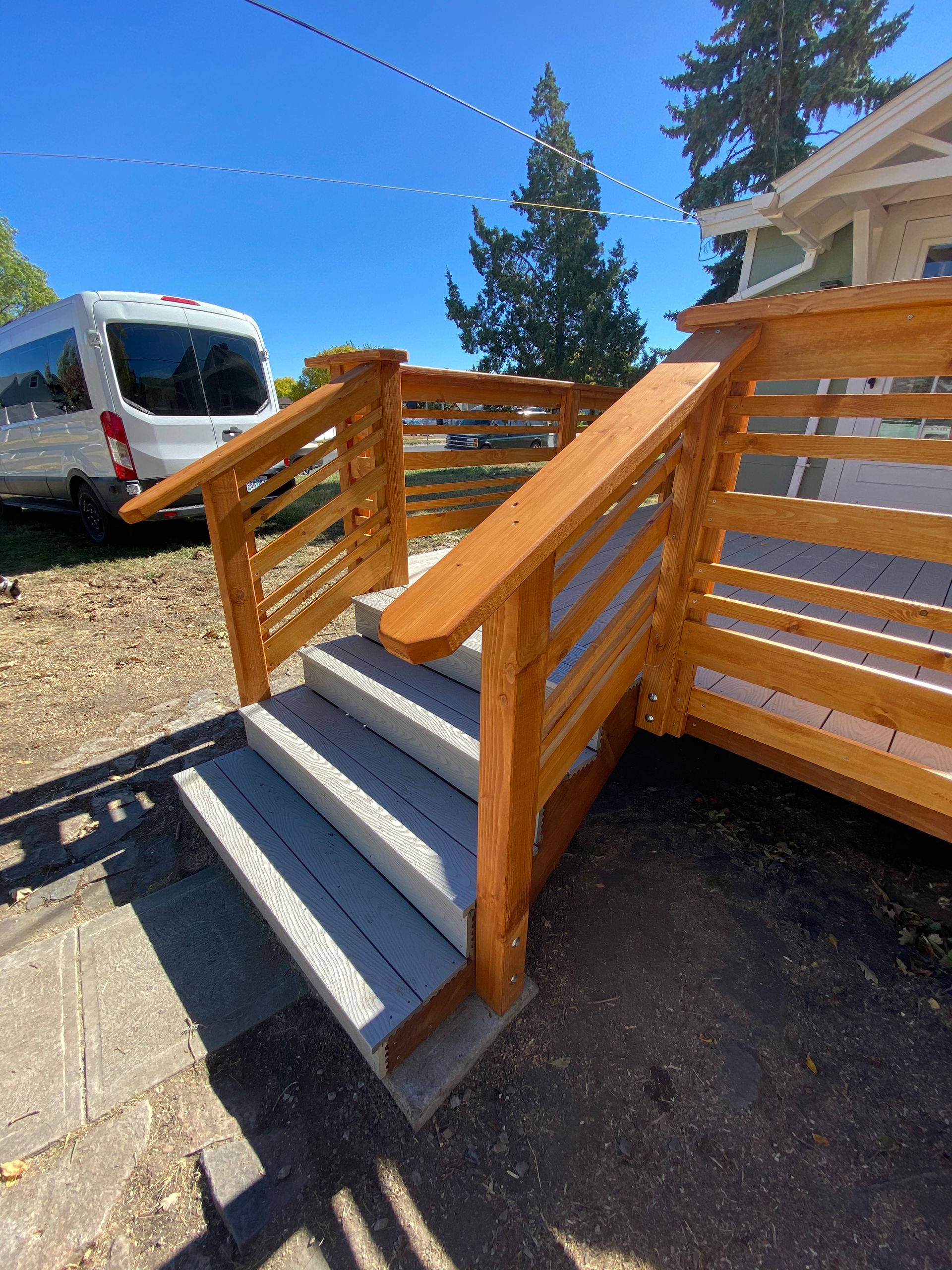 Outdoor wooden staircase with gray treads and railings leading up to a house exterior on a sunny day.