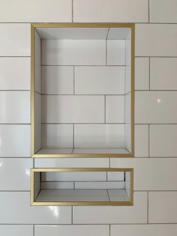 Two gold-trimmed rectangular recessed shower niches are set into a wall of white subway tiles.