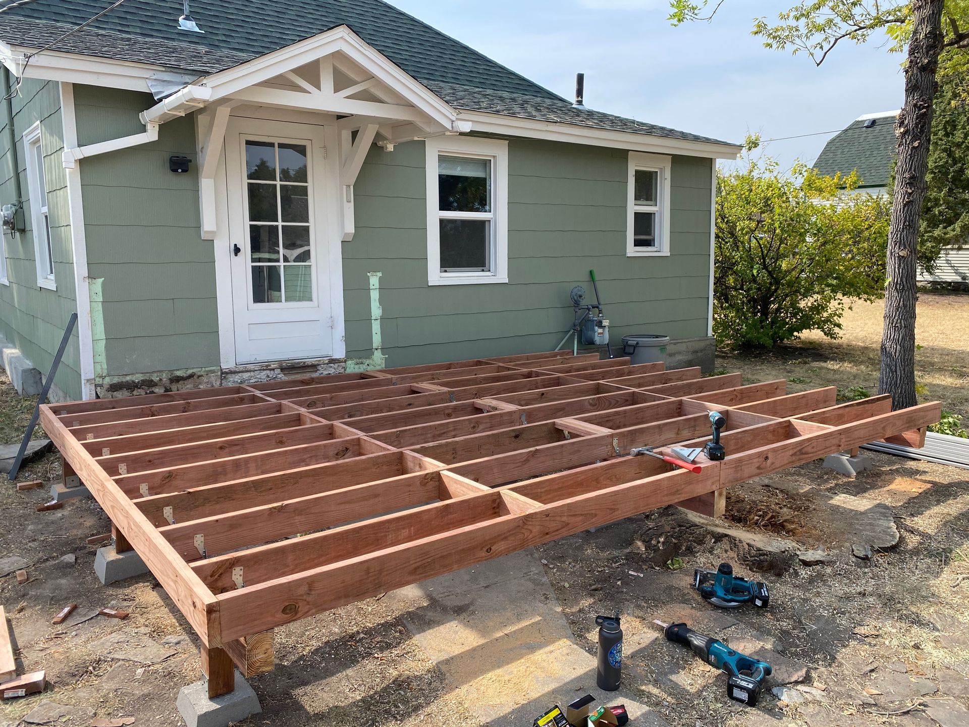 A wooden deck frame under construction in front of a green house, with power tools lying on the ground nearby.