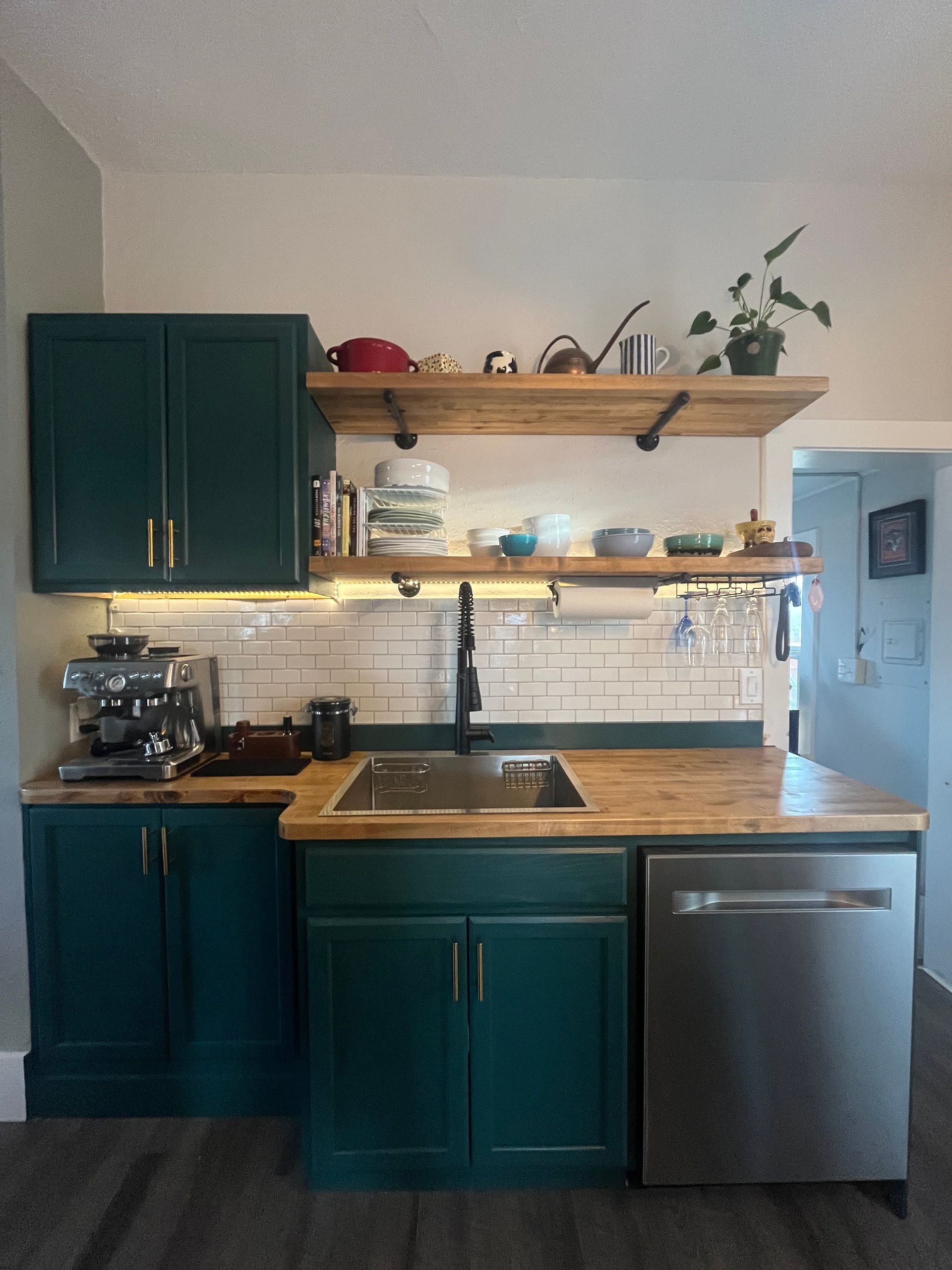 Dark teal kitchen cabinets with wooden countertops, open shelving, a subway tile backsplash, and stainless dishwasher.