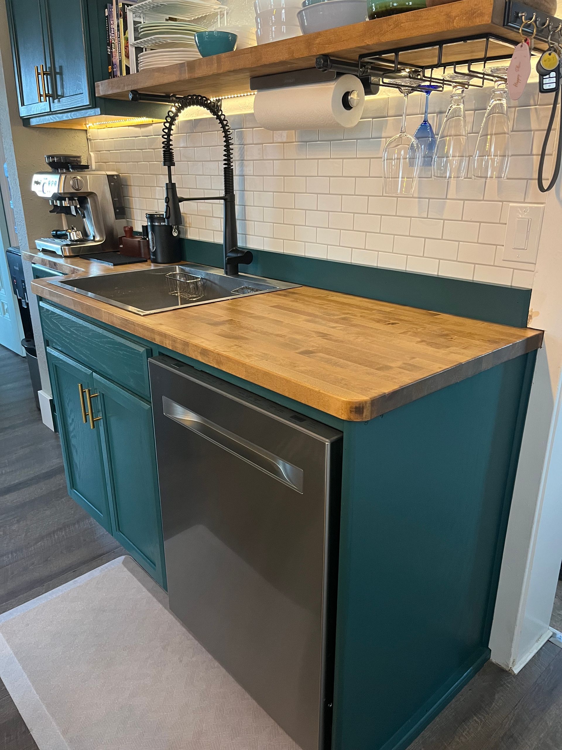 A kitchen area featuring a dark teal cabinet, wood countertop, stainless steel dishwasher, and a white tile backsplash.