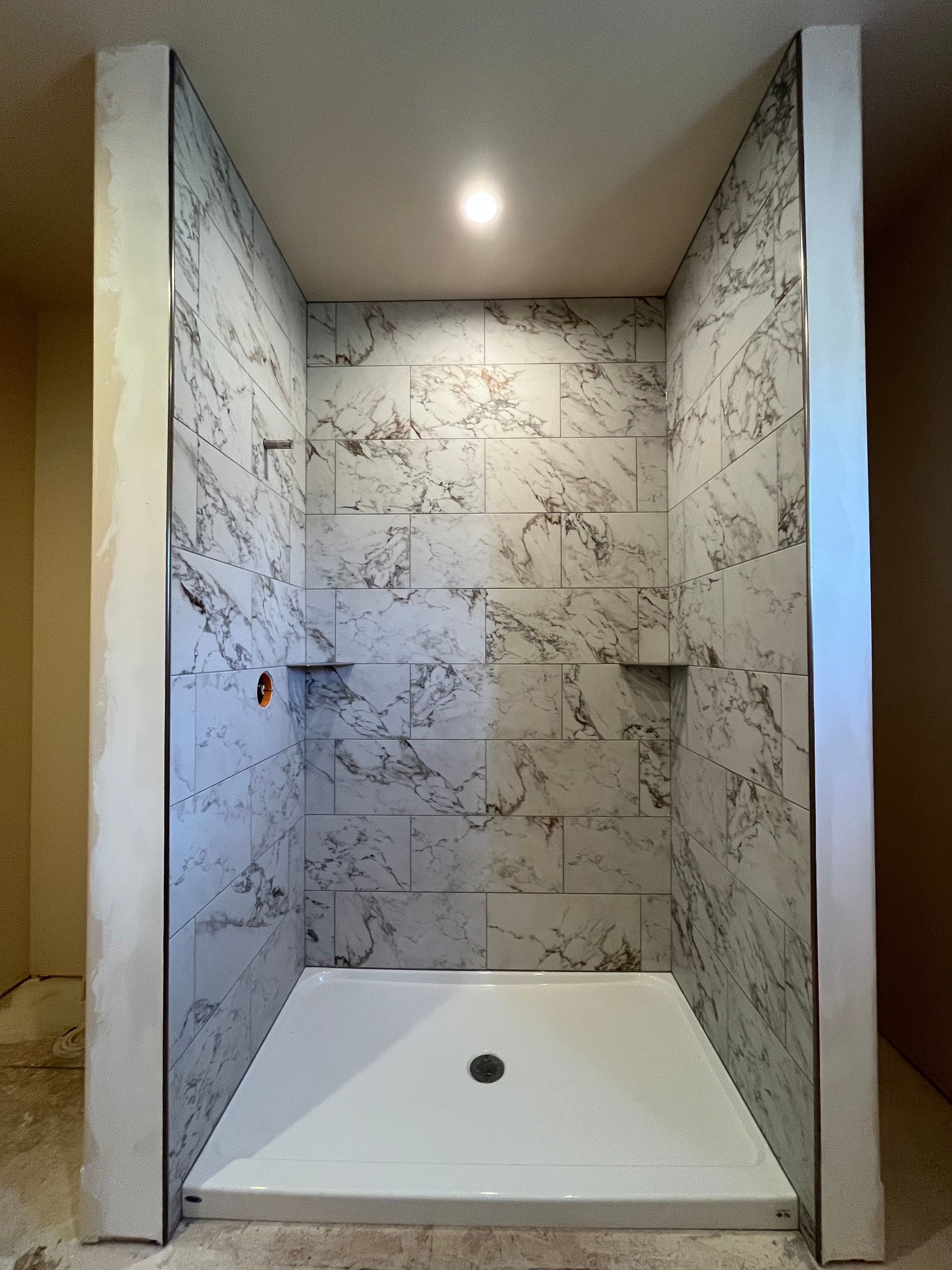 A walk-in shower with white, grey-veined marble-patterned tiles, a white shower pan, and silver trim under a spotlight.