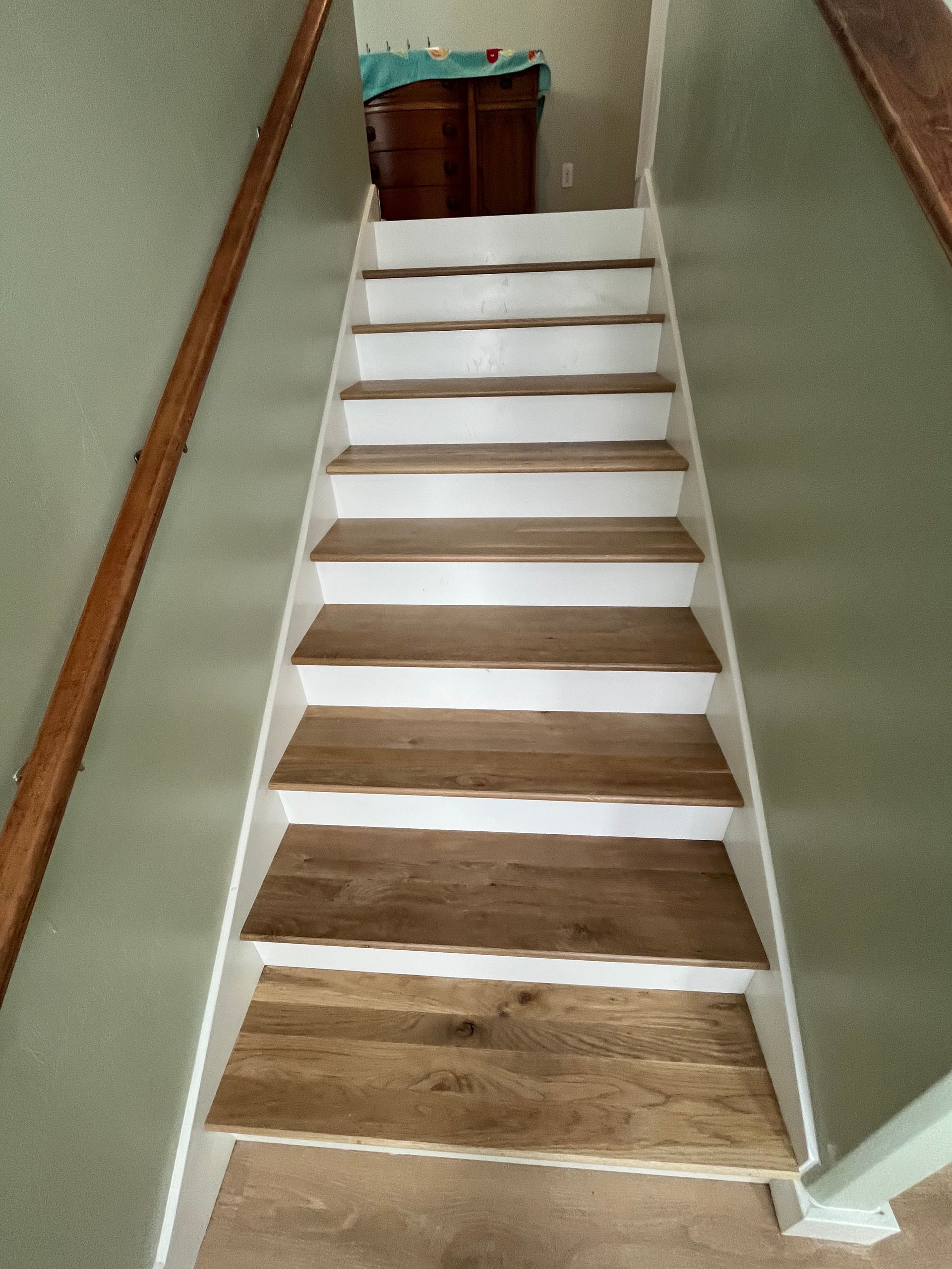 A straight set of indoor stairs with light wood treads and white risers, framed by green walls and wooden handrails.