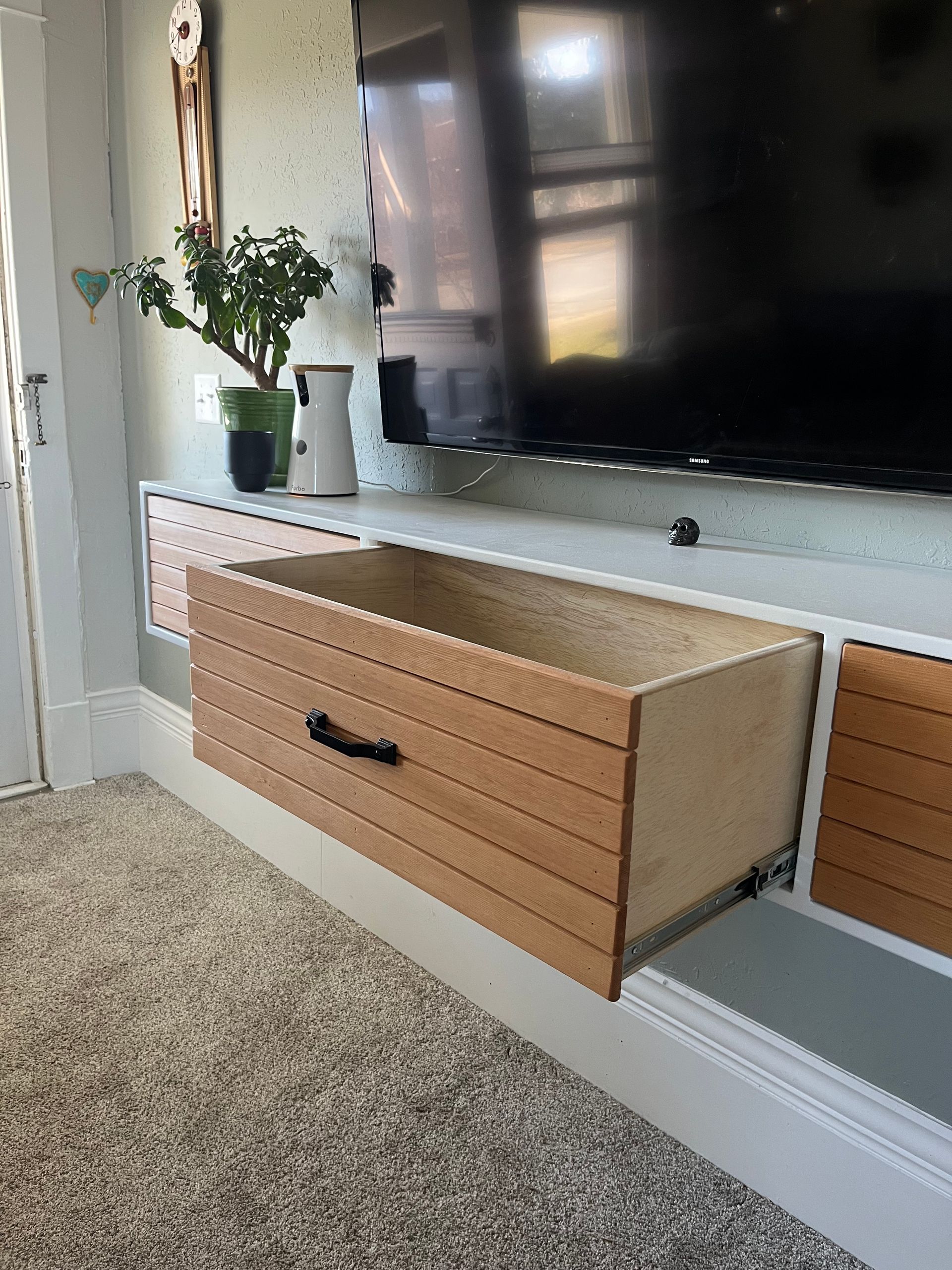 A wooden drawer, featuring horizontal slat details and a black handle, pulled open from a wall-mounted white media unit.
