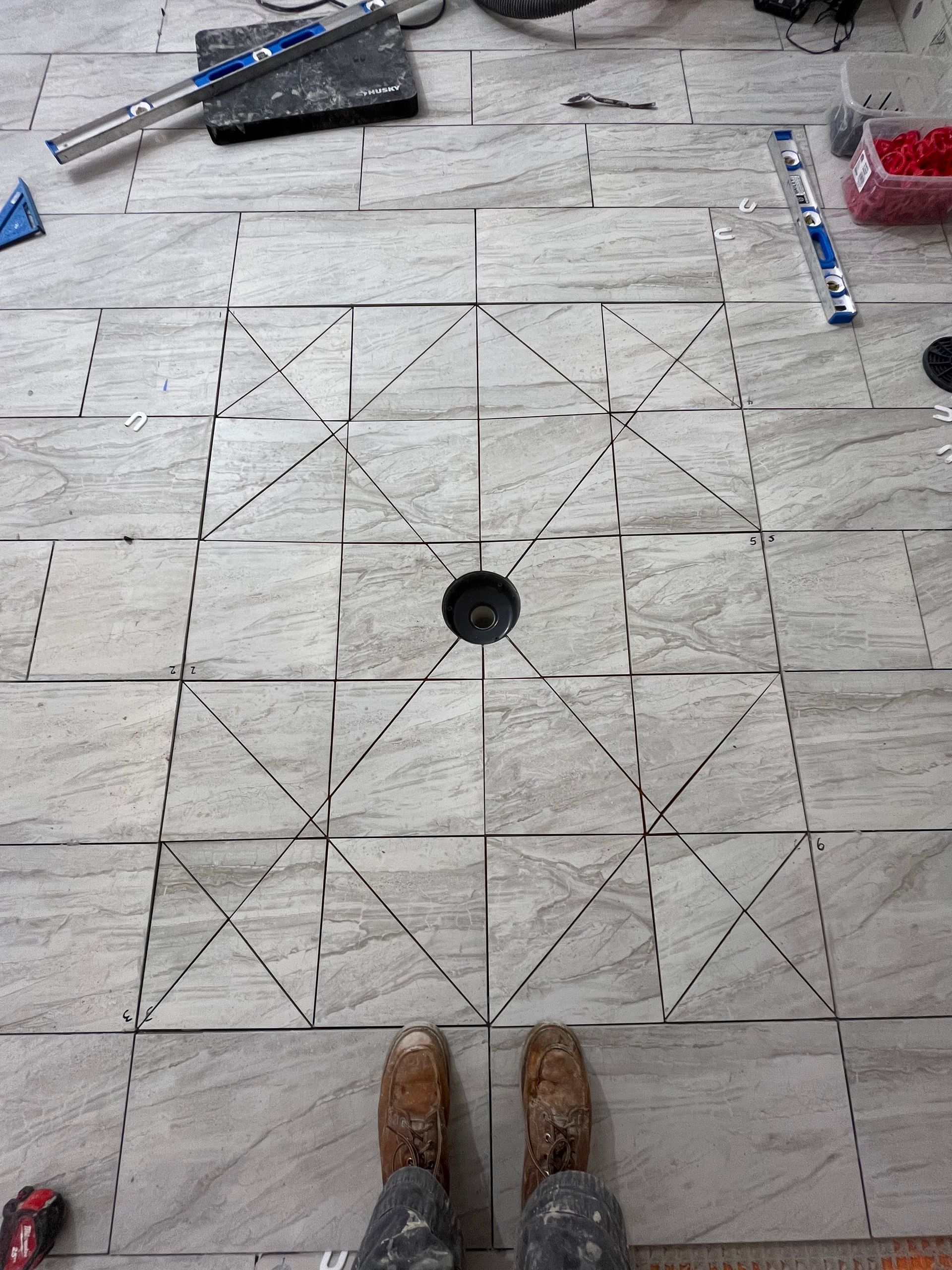 A person stands on floor tiles being installed around a central drain, showing diagonal cuts for a shower pan slope.
