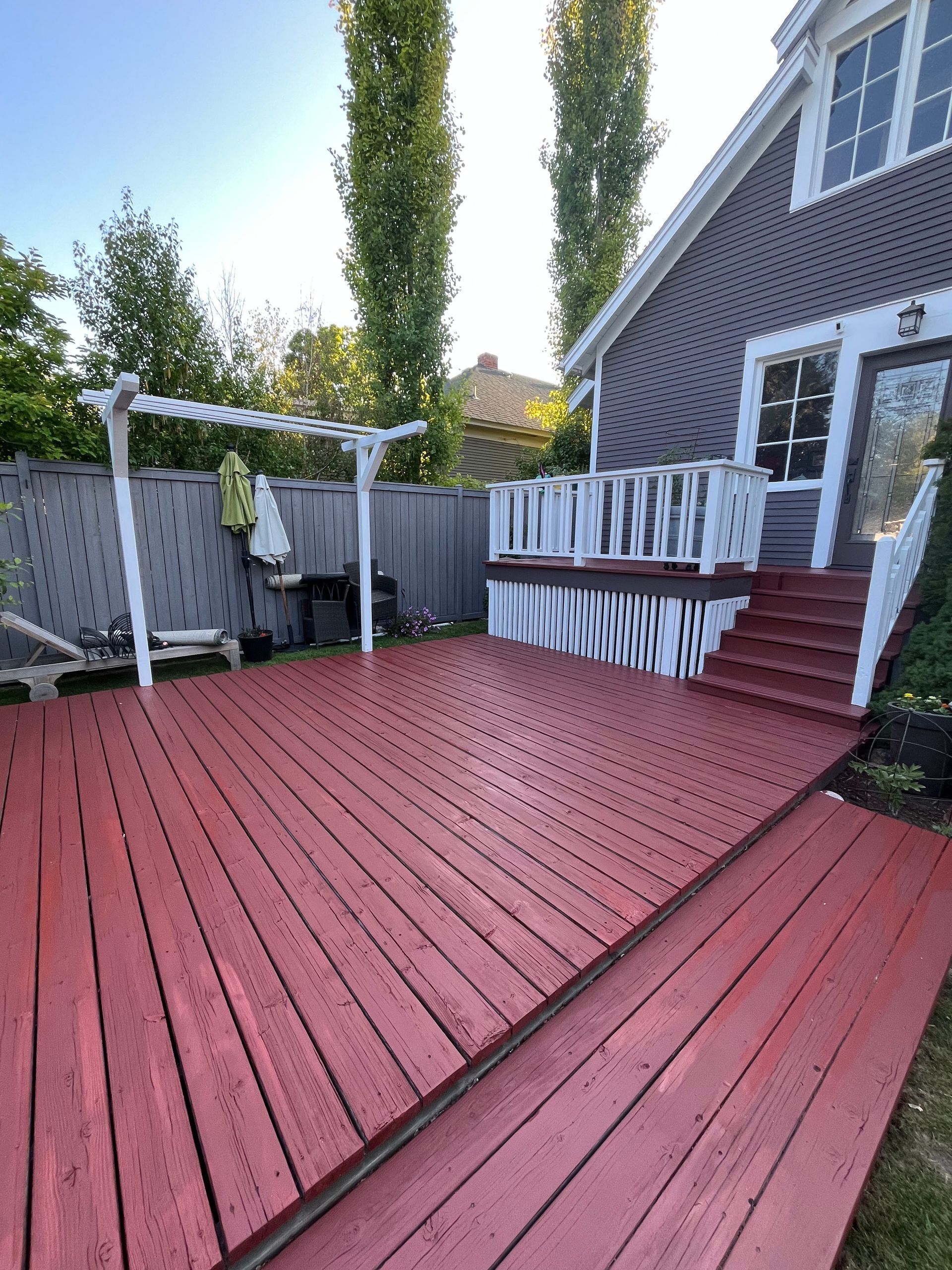 A large, red wooden deck with a white trellis, white railing, and stairs leading to a grey house exterior.