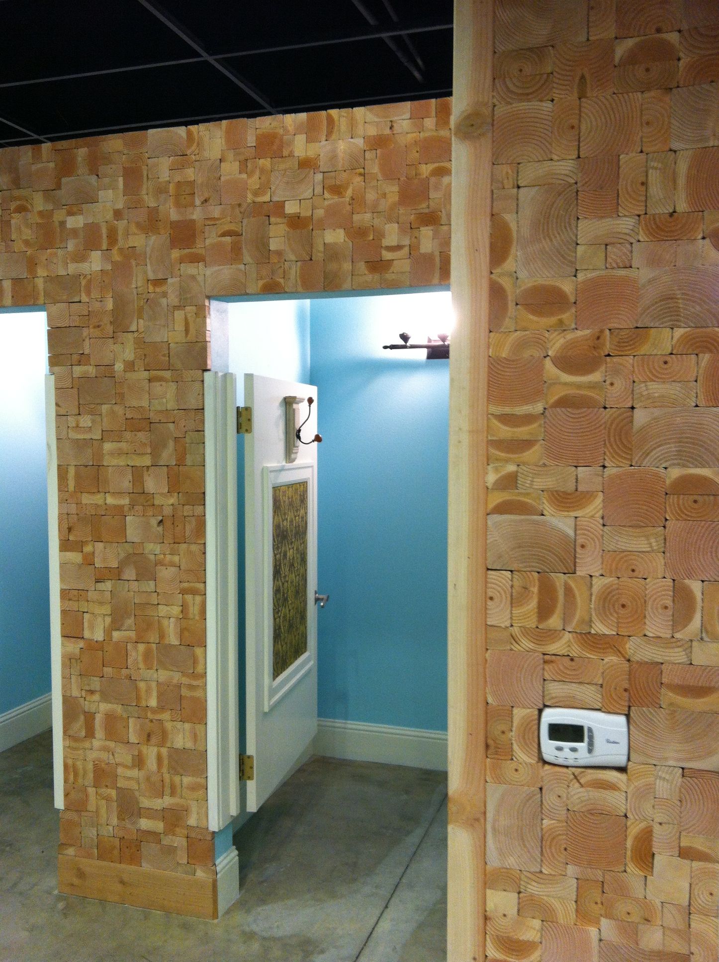 A doorway leads into a blue-walled restroom, framed by walls covered in small, textured, light brown wood block tiles.