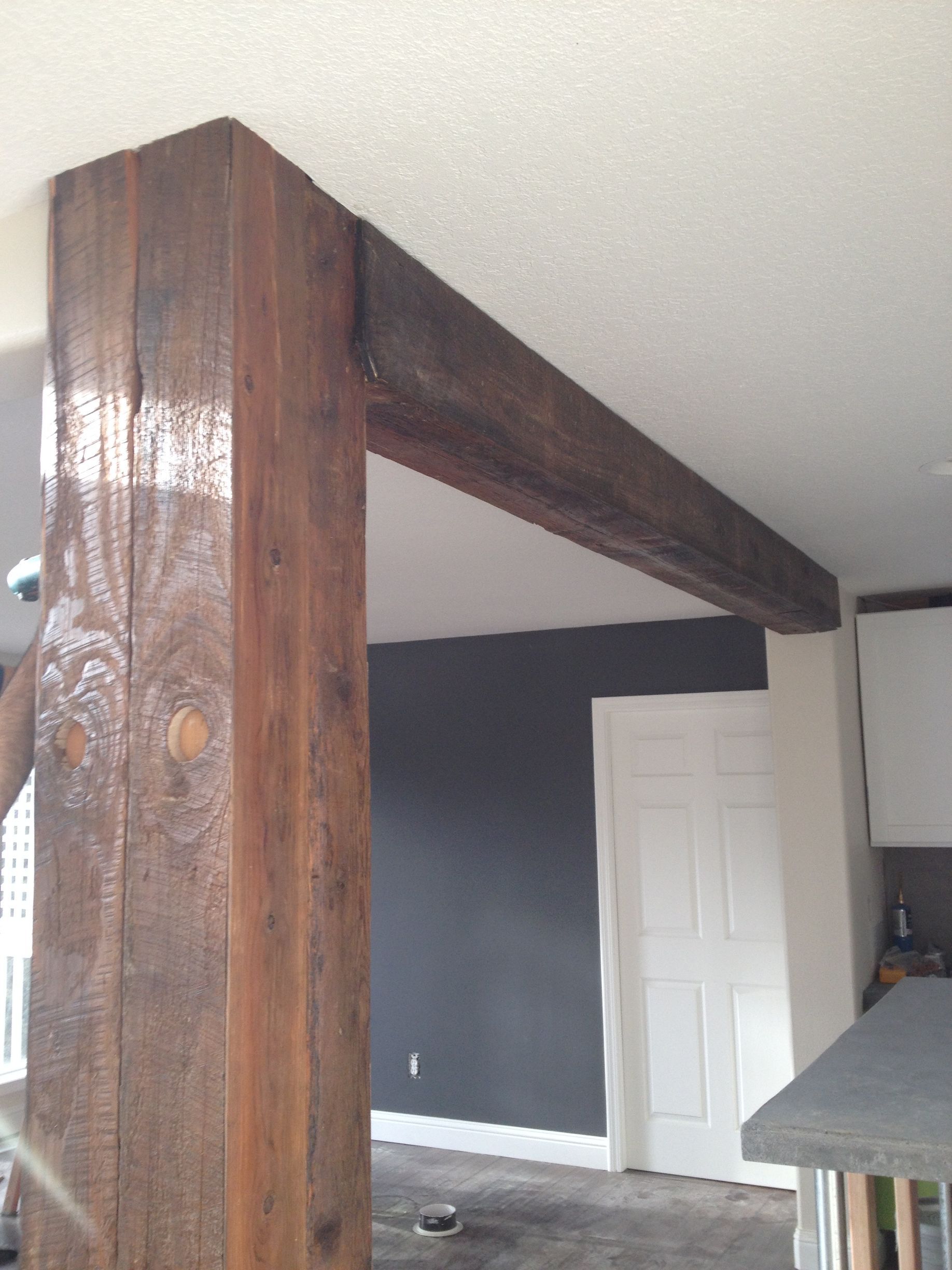 A wooden post and ceiling beam finished with a dark stain in a home interior with a gray wall and white door in the back.