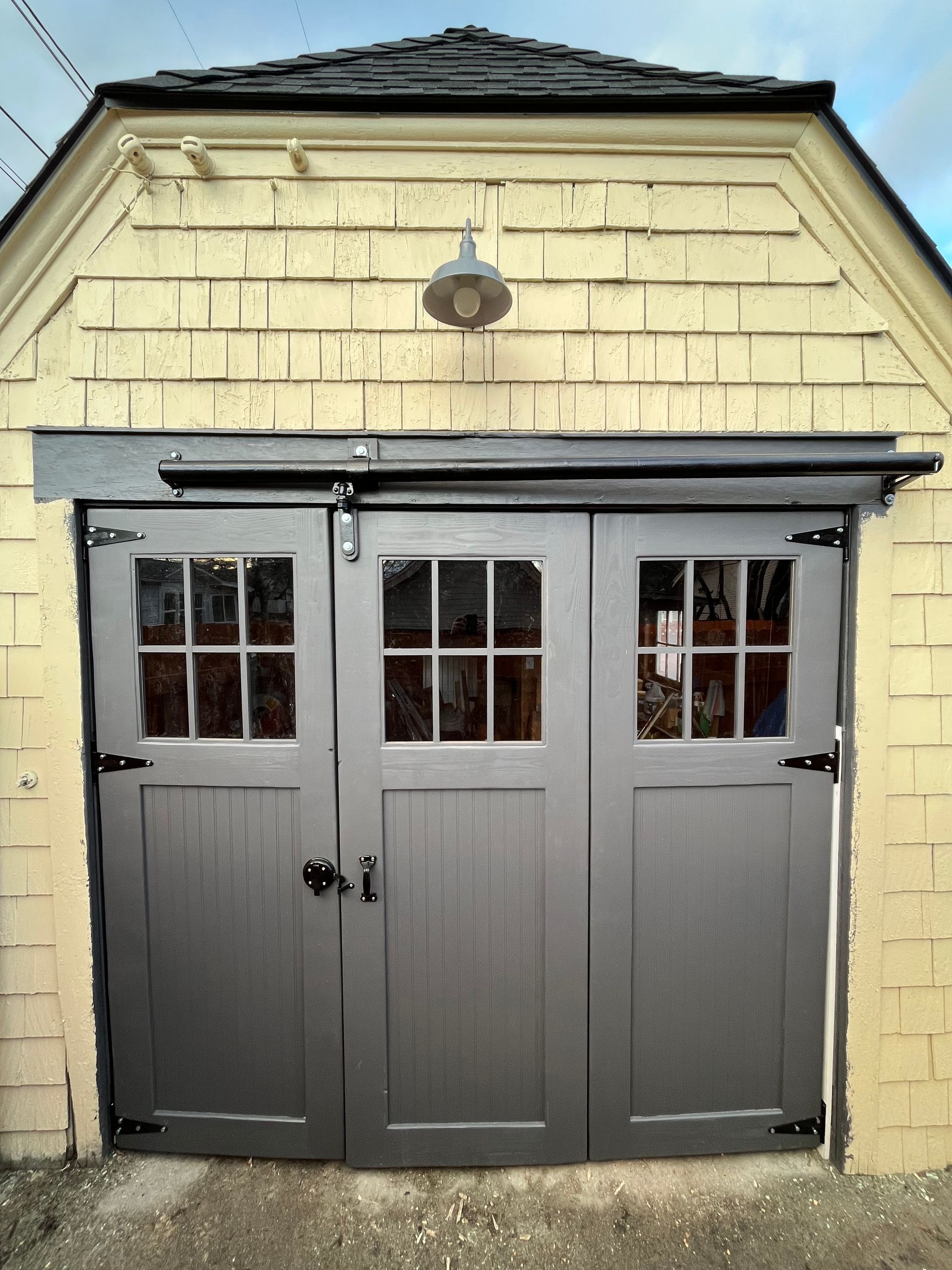 Dark grey, three-panel garage doors with windows, set in a light yellow, shingled building exterior with a wall light.