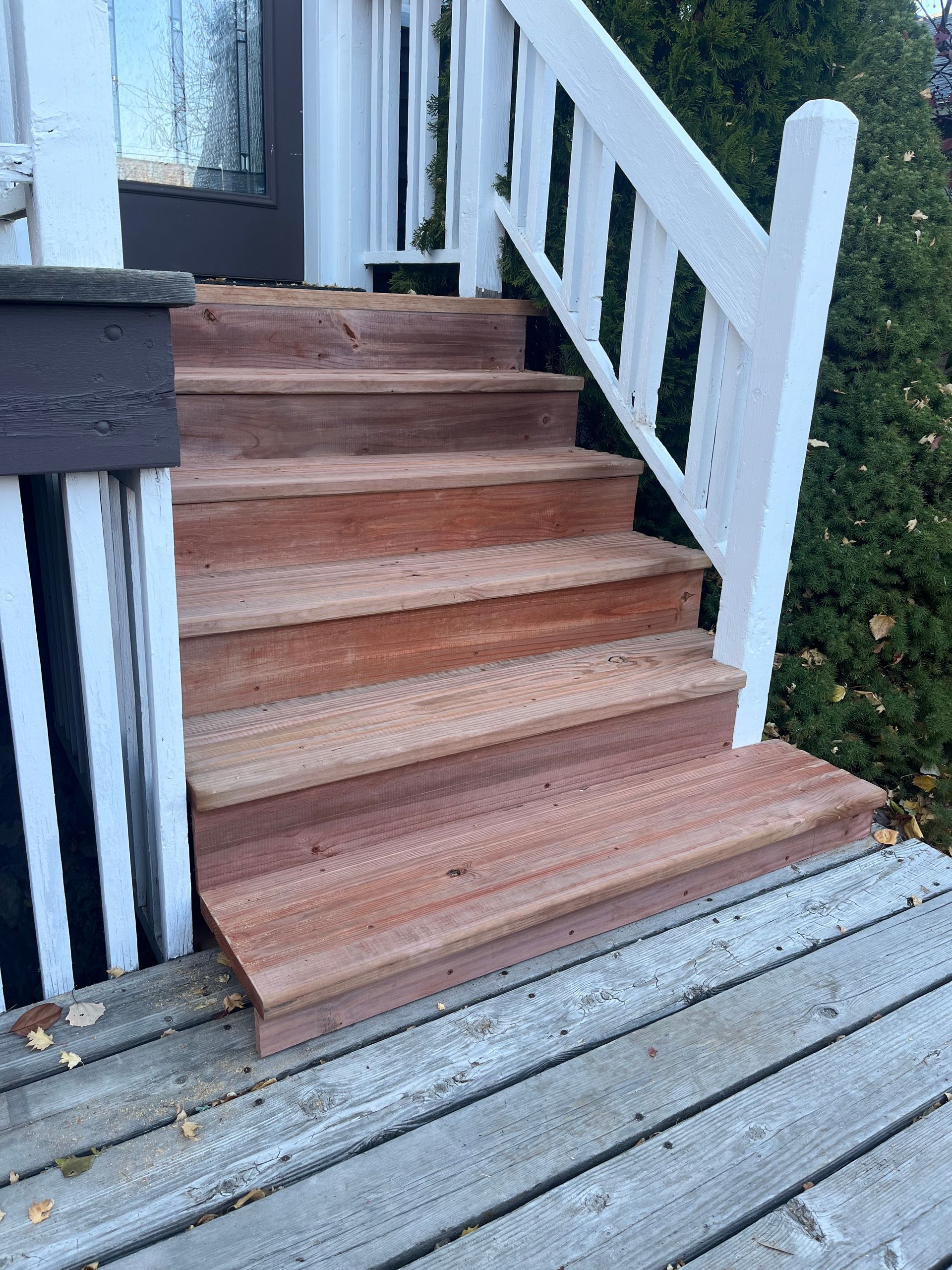 A set of five natural wood steps leading up to a porch with white railings and a gray wooden deck.