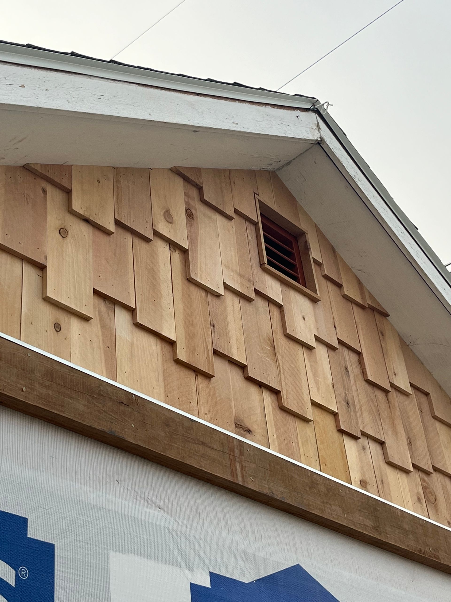 Wooden shingles installed on a gable end, featuring a small square vent near the roofline above a white house wrap barrier.