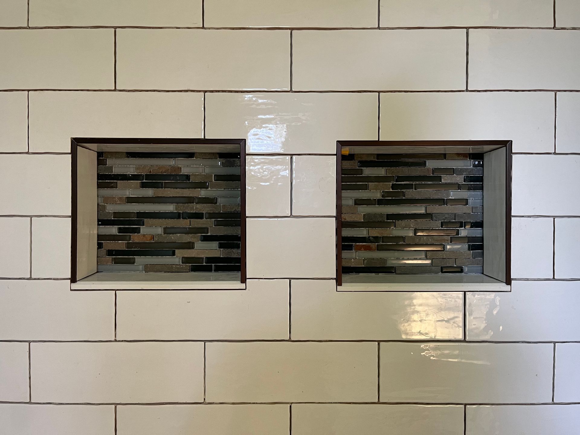 Two side-by-side recessed shower niches tiled with dark, horizontal glass mosaic set into a white subway tile wall.