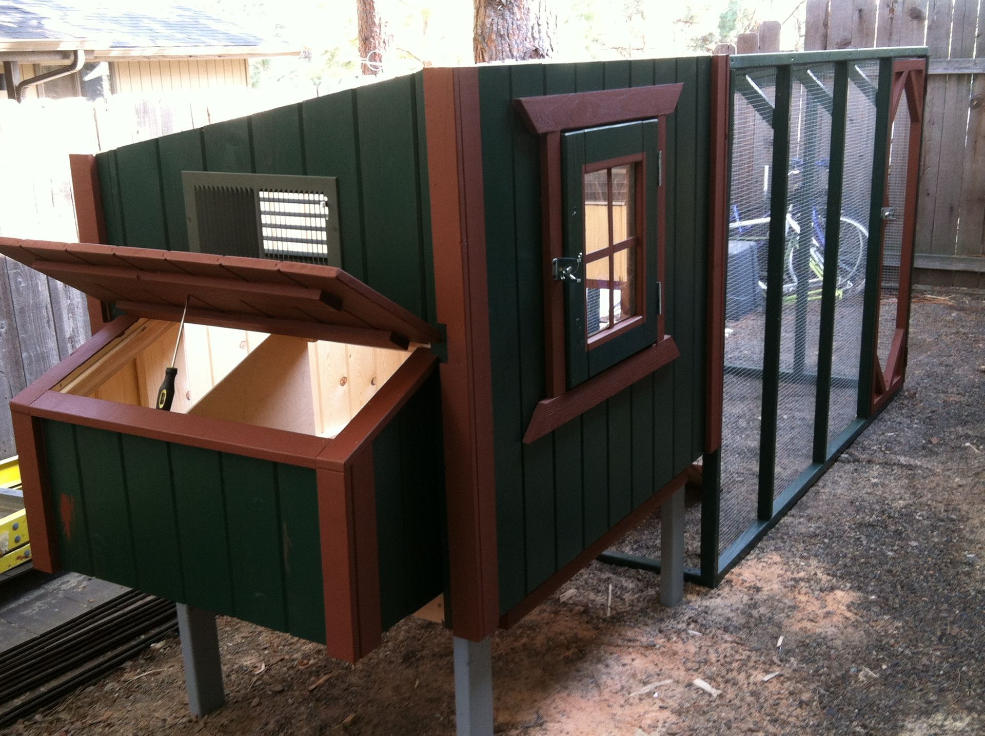 A dark green and brown wooden chicken coop stands on legs in a yard, with an open nesting box and an attached wire run.