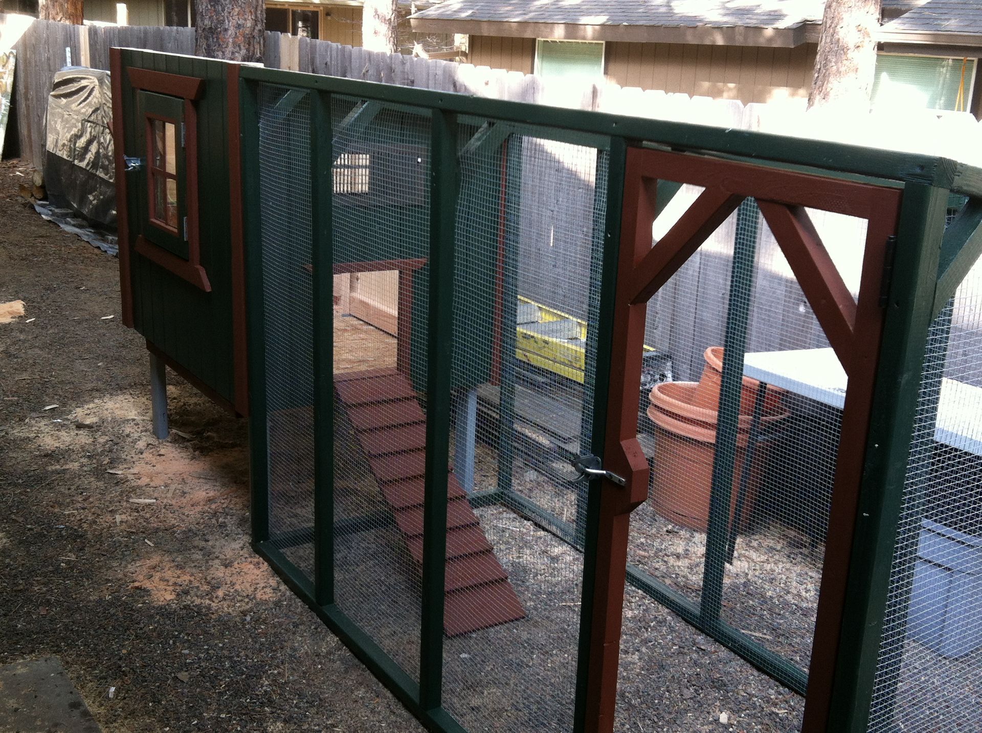 A green and brown wooden chicken coop with a mesh wire run, a ramp, and a small enclosed house in a backyard.