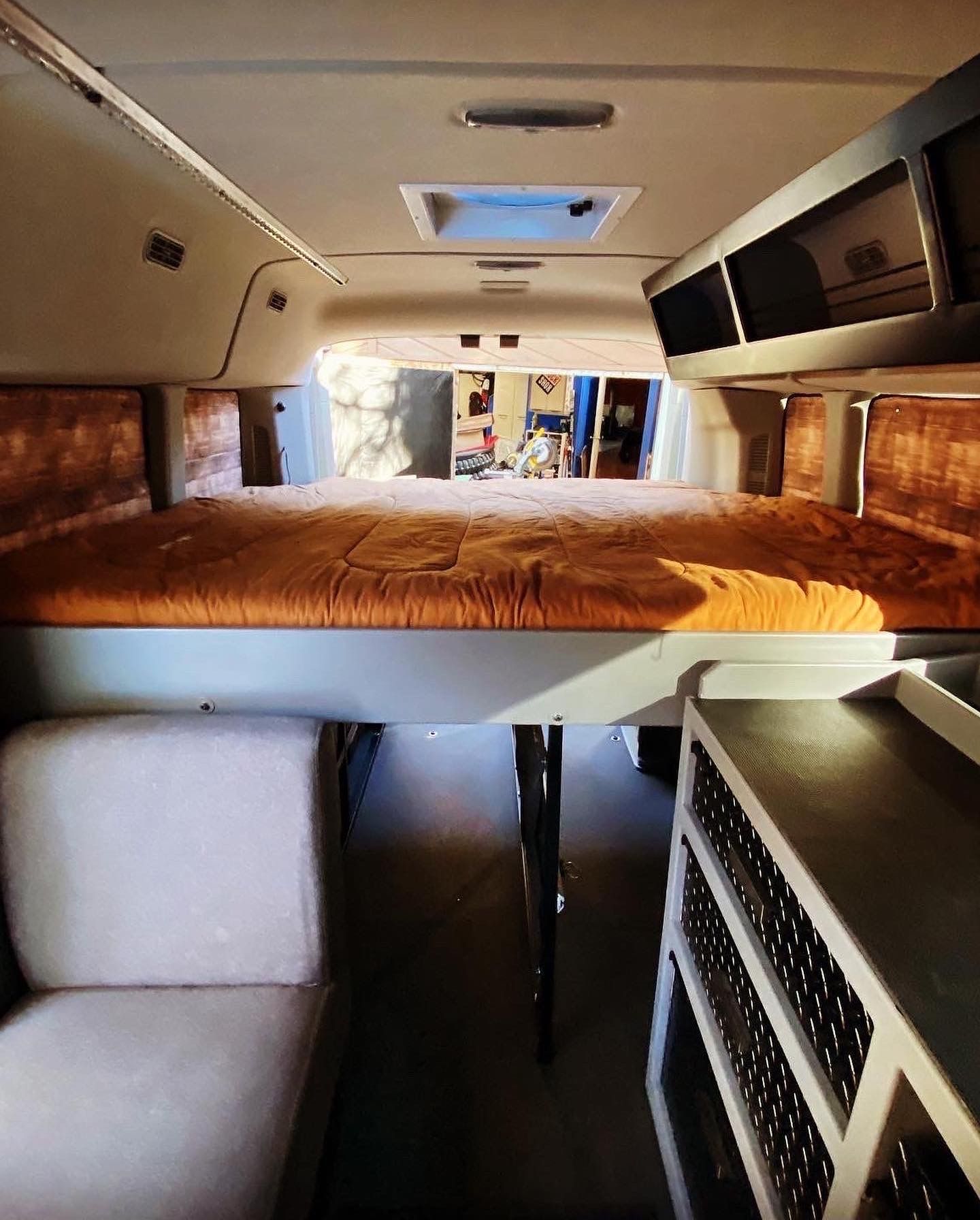 Interior view of a camper van featuring a raised platform bed with an orange mattress, a small seat, and storage units.